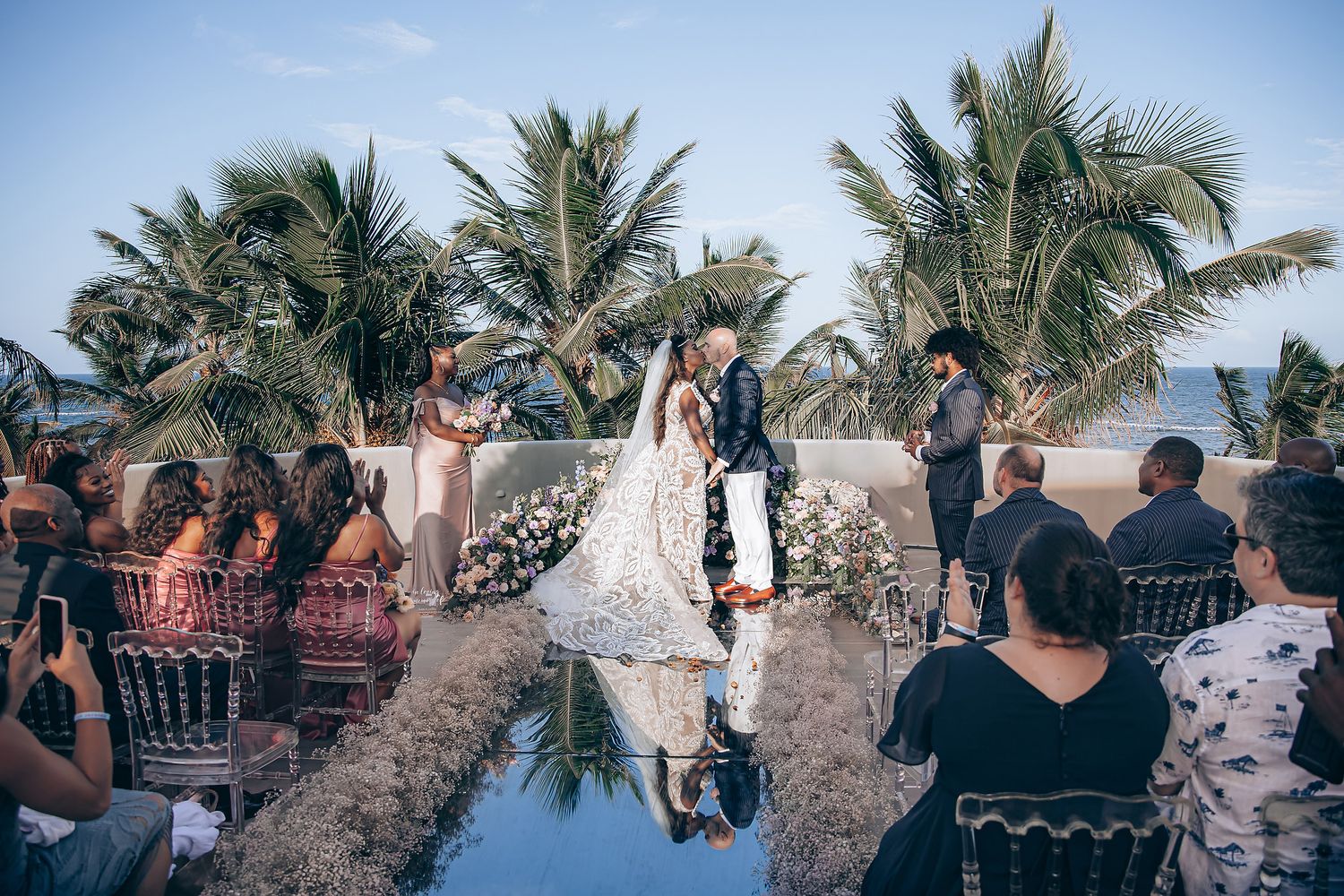 Wedding guests gather under palm trees at a tropical beach ceremony during sunset.