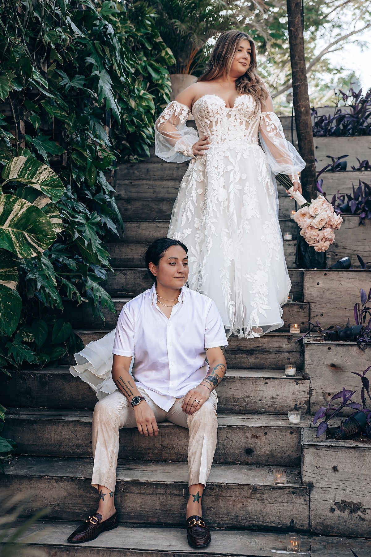 Couple in white outfits poses on rustic wooden steps surrounded by greenery during an outdoor photoshoot.