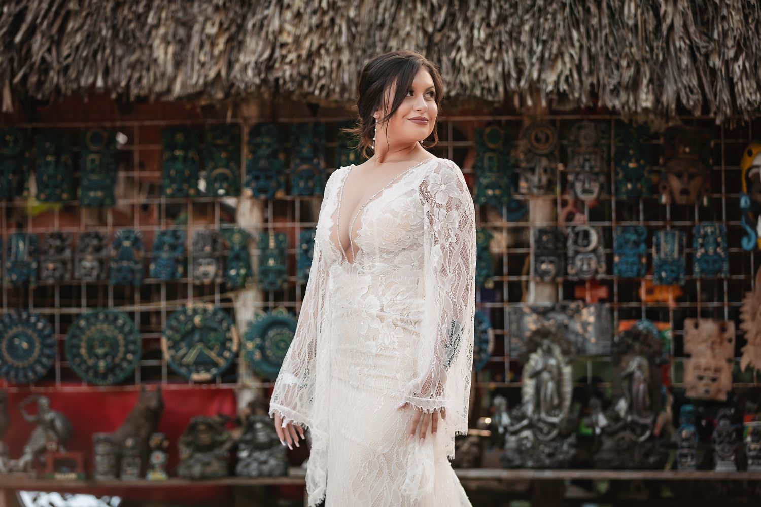 A stunning lace wedding dress with long sleeves and intricate details photographed under a thatched roof structure.
