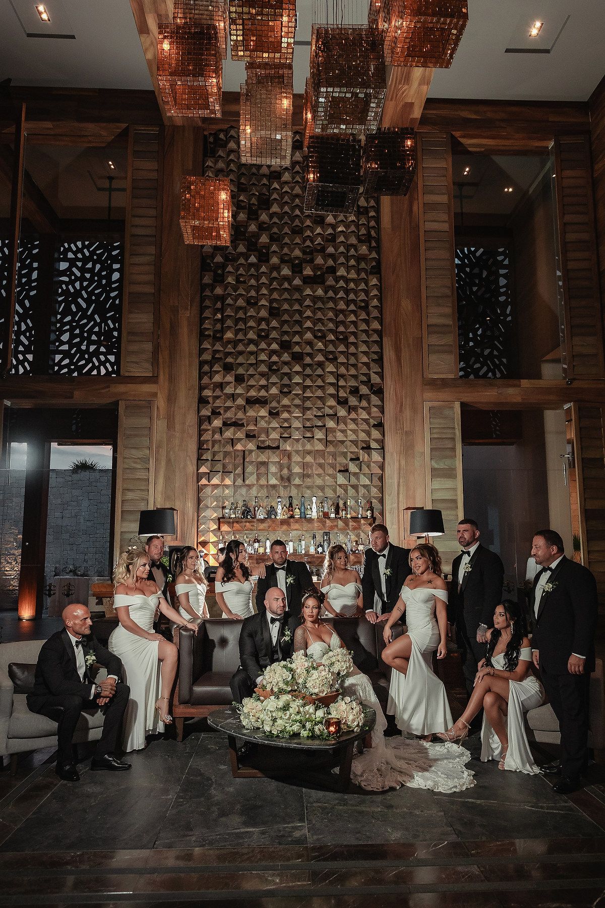 Wedding party poses together in front of brick wall with romantic copper lighting and modern decor.