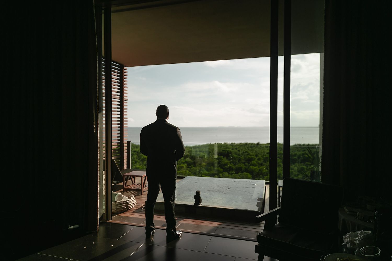 A silhouetted figure stands at a window overlooking a scenic coastal landscape during sunset.