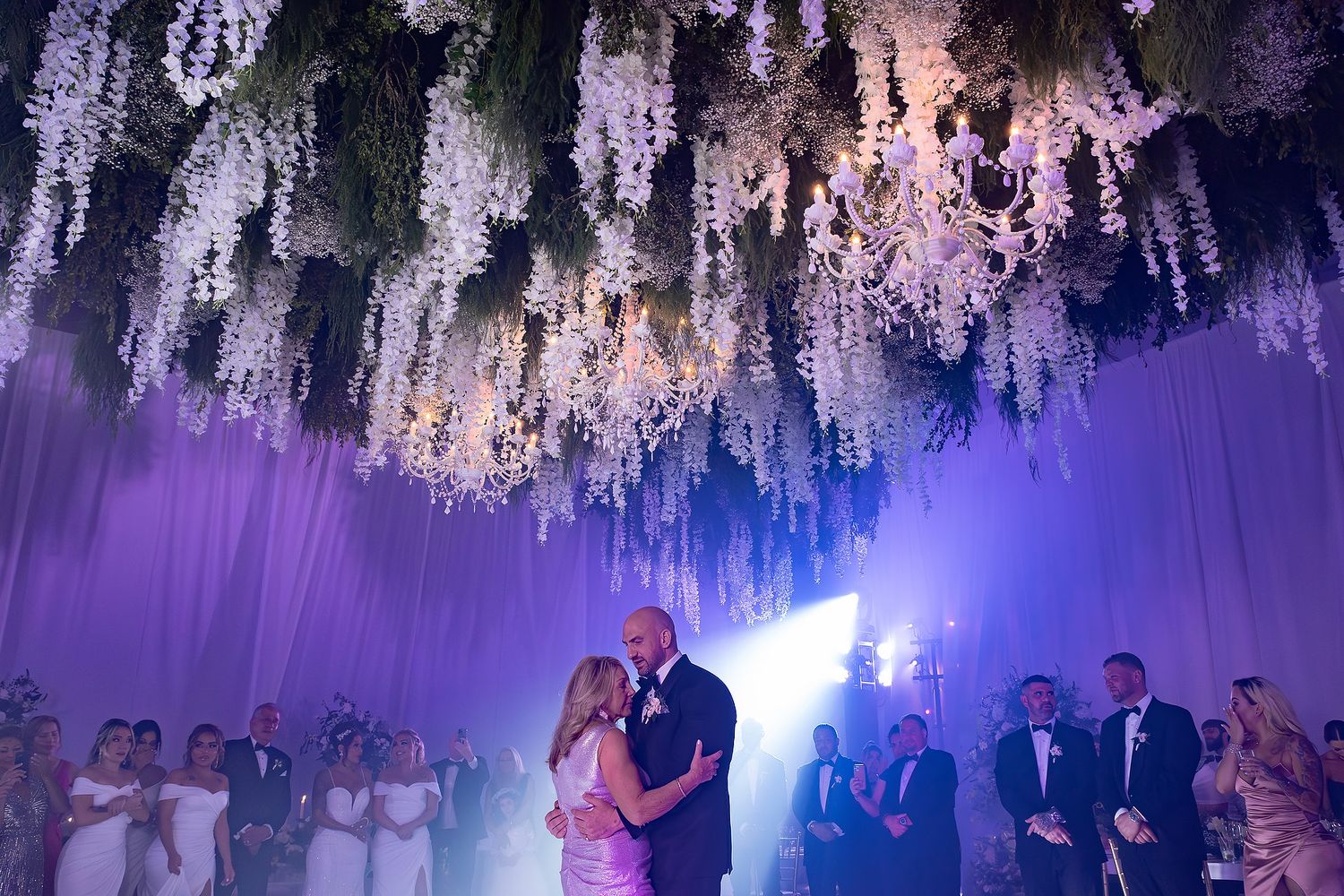 Elegant wedding reception with cascading white floral ceiling decorations illuminated by dramatic purple and blue lighting.