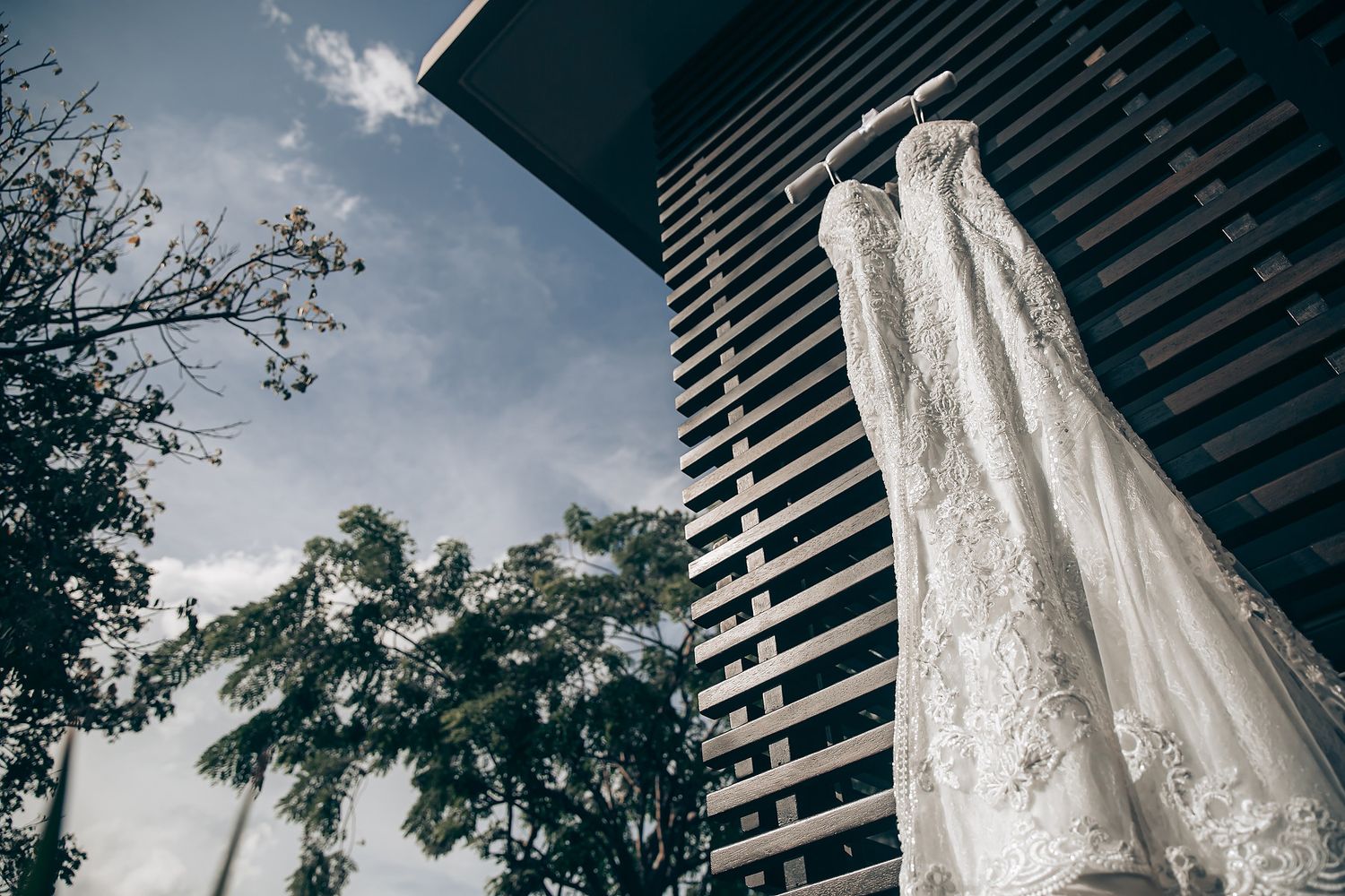 Elegant white lace wedding dress hanging outdoors against modern architecture and greenery.