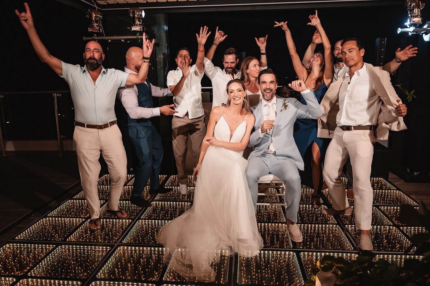 Celebratory group dance photo with wedding party on illuminated floor during evening reception.