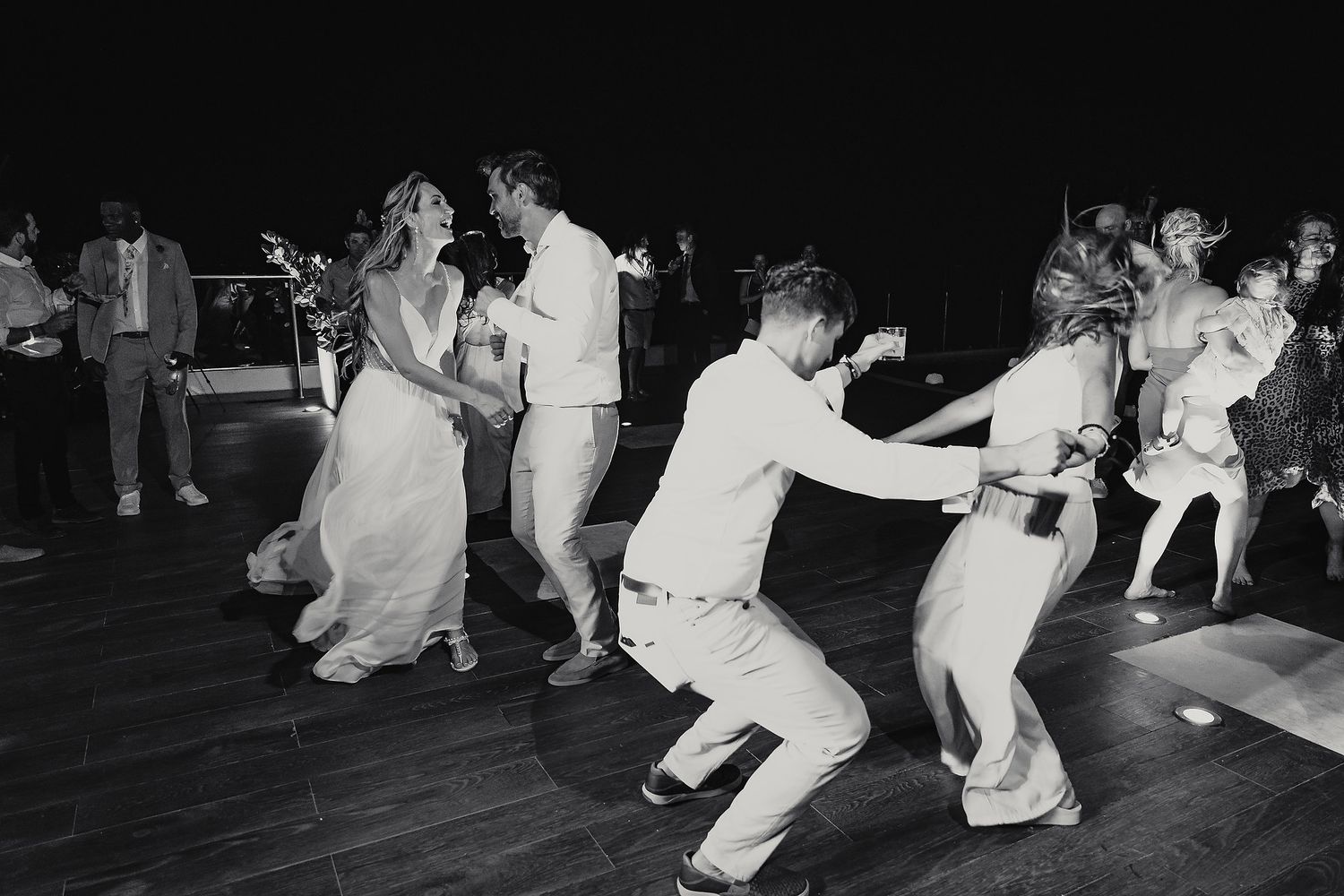 People in white outfits dance in formation on a dimly lit dance floor at a celebration.