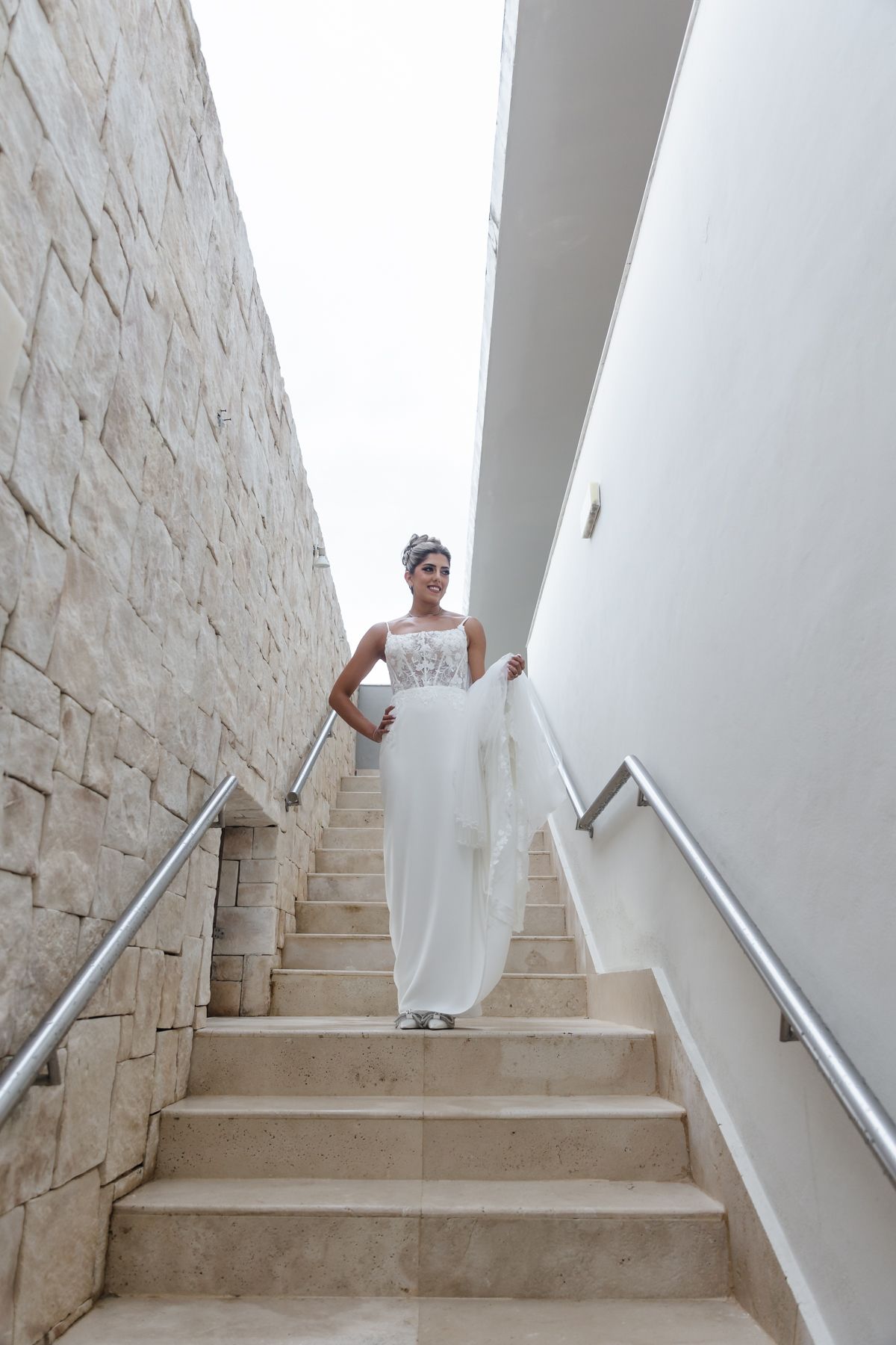 Bride in white gown descending stone stairs between brick and white walls, holding her flowing dress train.