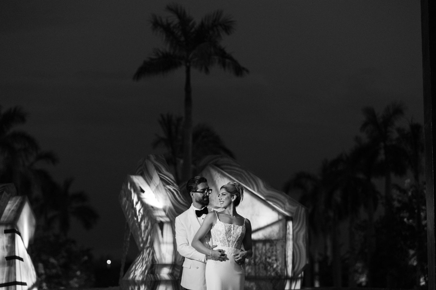 Black and white wedding portrait of couple embracing under palm trees at night.