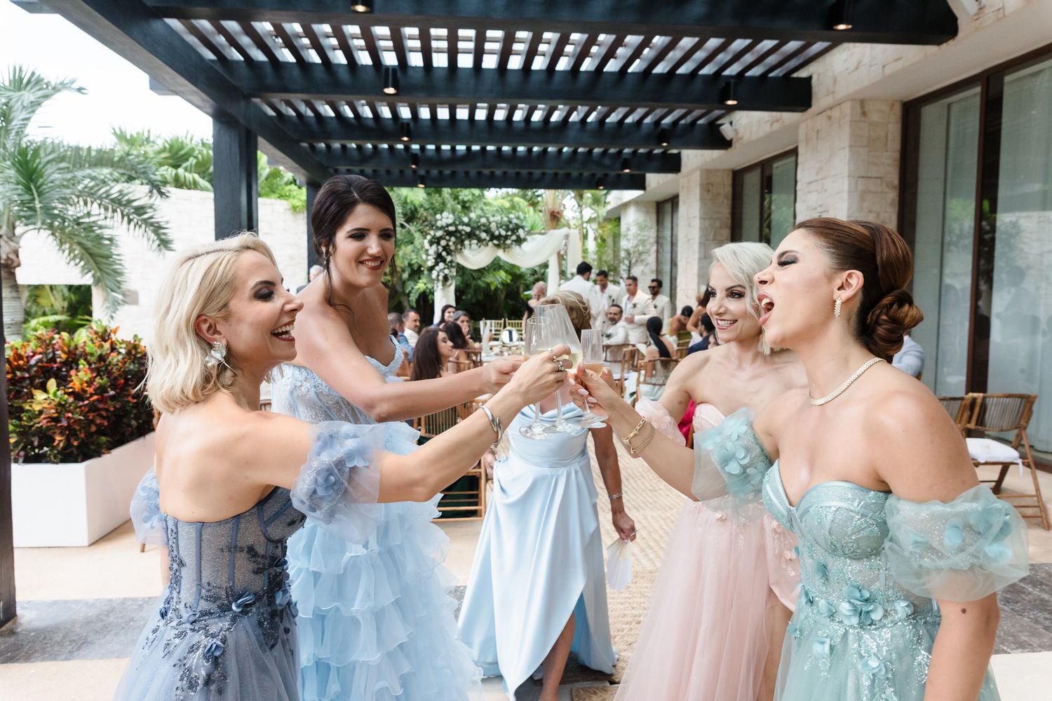 Bridesmaids in blue off-shoulder gowns toasting champagne glasses outdoors at a tropical wedding celebration.