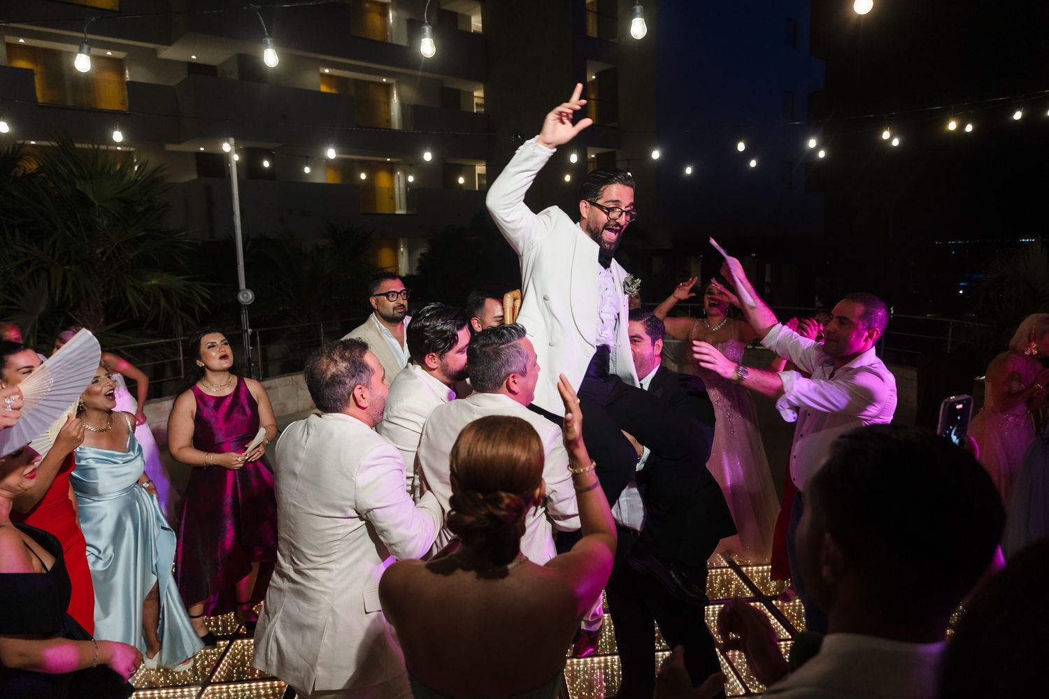 Groom lifted by guests at outdoor wedding reception with string lights and festive crowd.