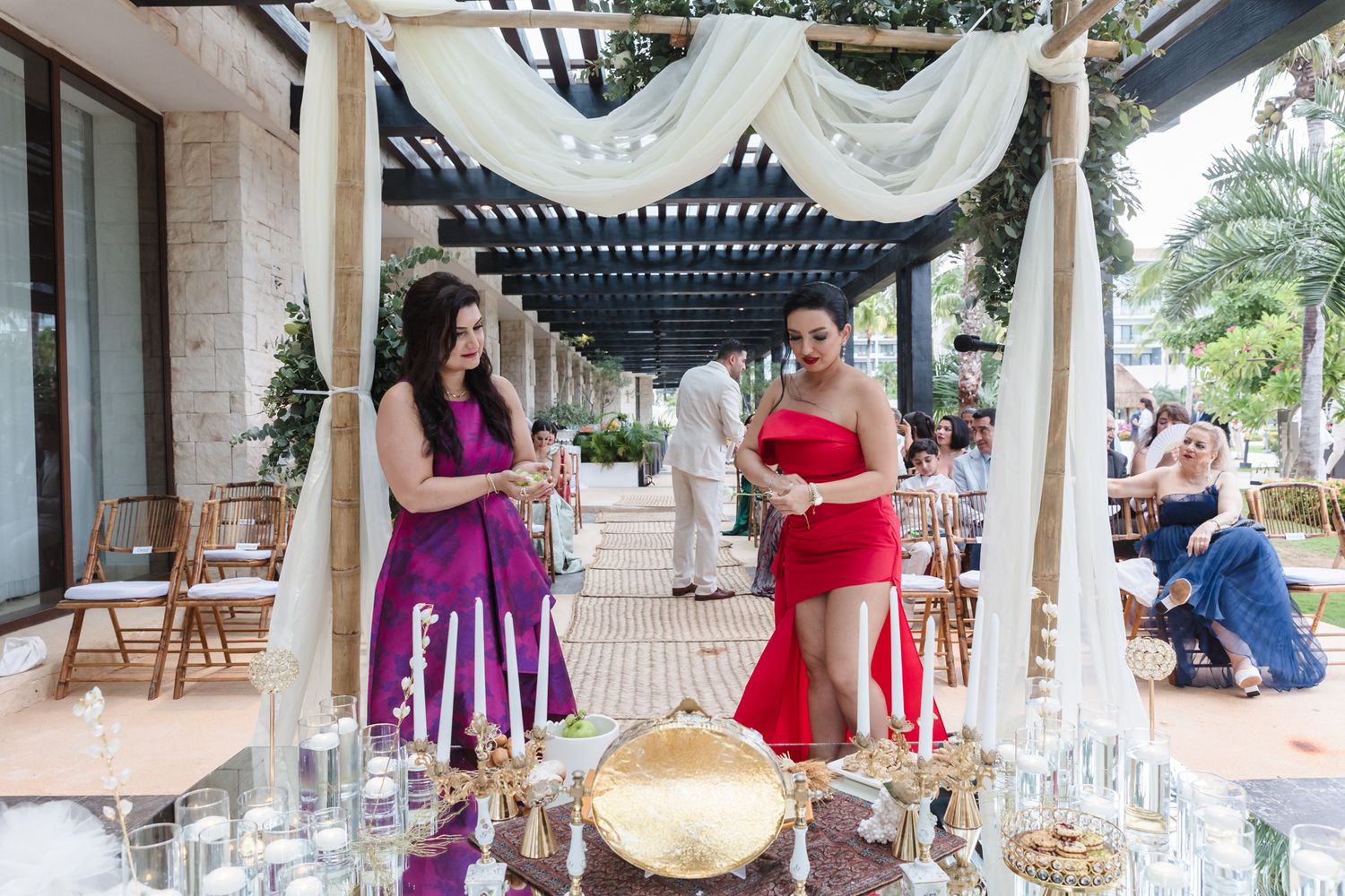 Two women in colorful dresses stand at an elegantly decorated outdoor wedding ceremony with candles and floral arrangements.