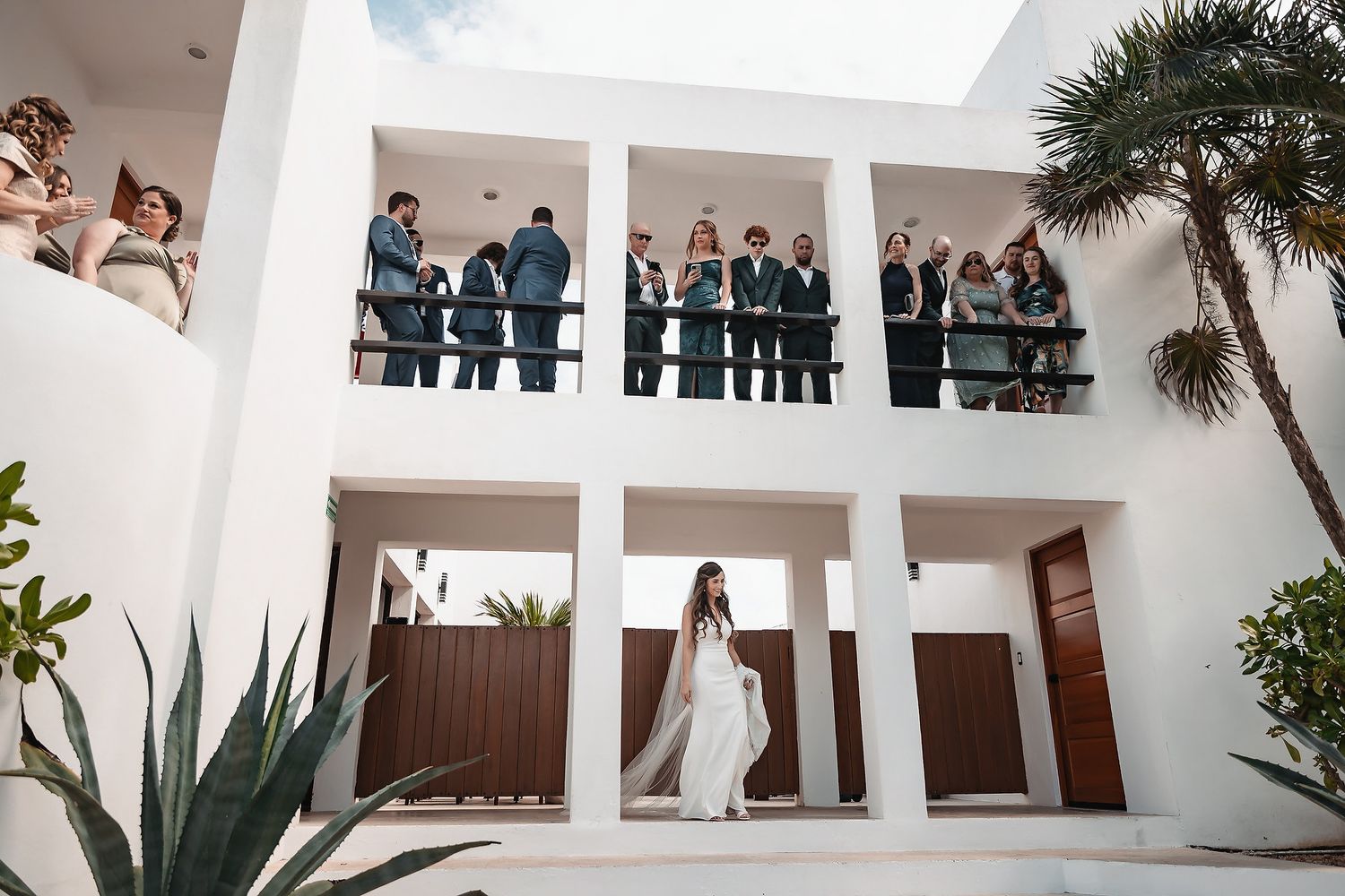 Bride in white gown stands beneath balcony where wedding guests gather at a modern white venue.