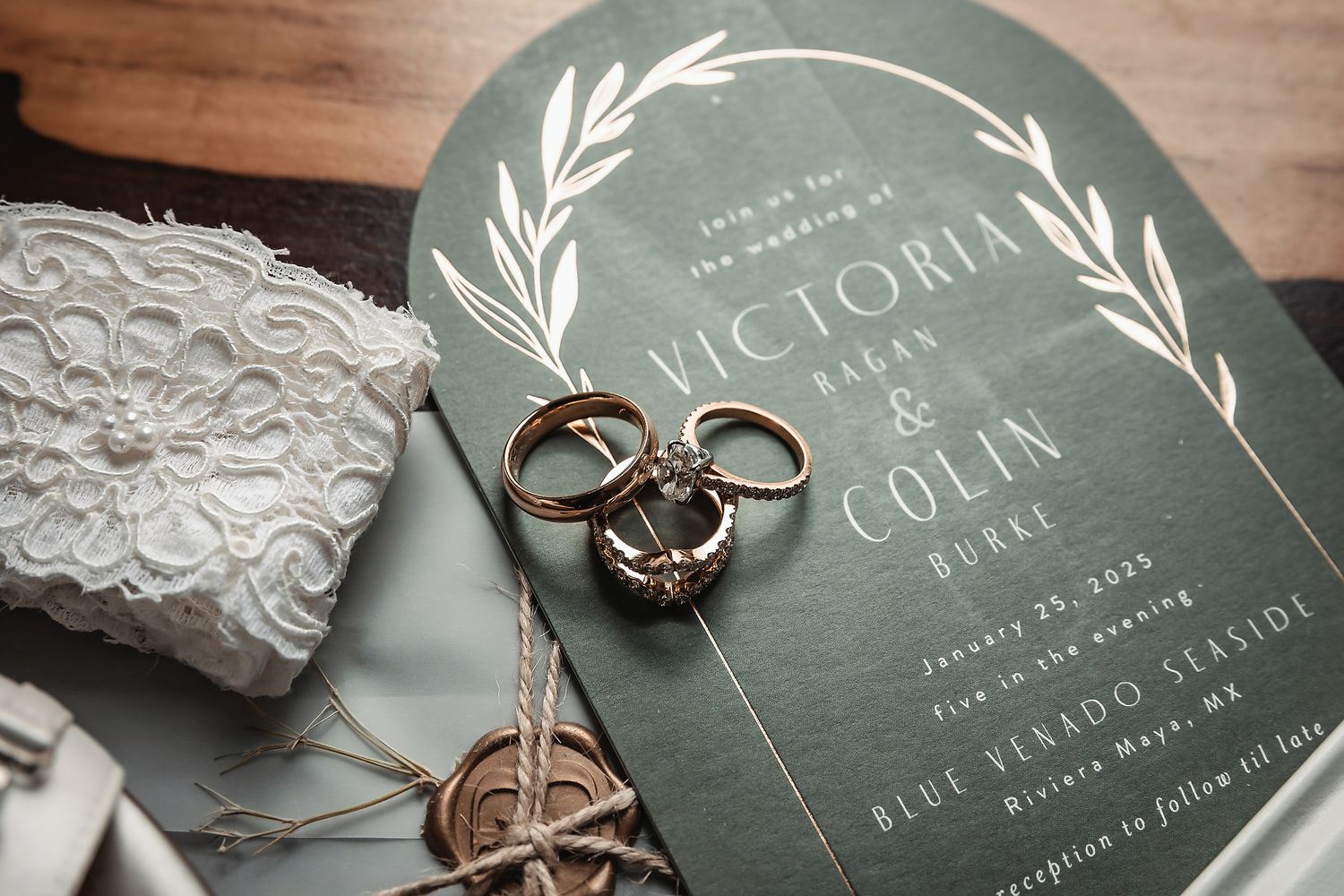 Green wedding invitation for Victoria and Colin with gold rings and lace details on a wooden surface.