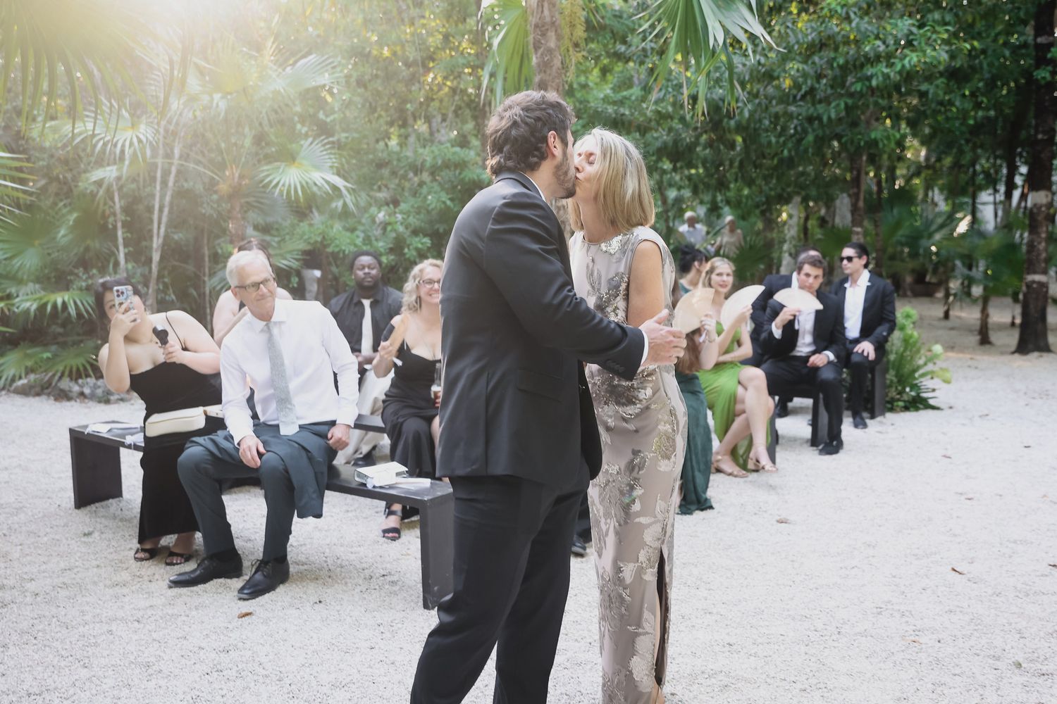 Couple shares a kiss during outdoor wedding ceremony surrounded by seated guests amid lush tropical trees.
