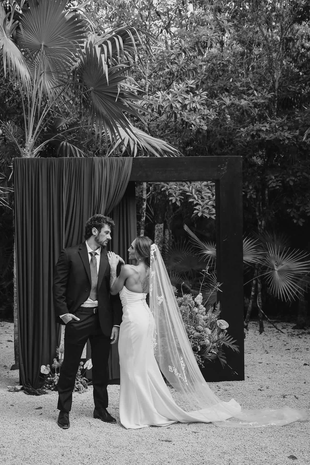 Black and white photo of bride and groom standing before a dark floral arch surrounded by tropical palms.