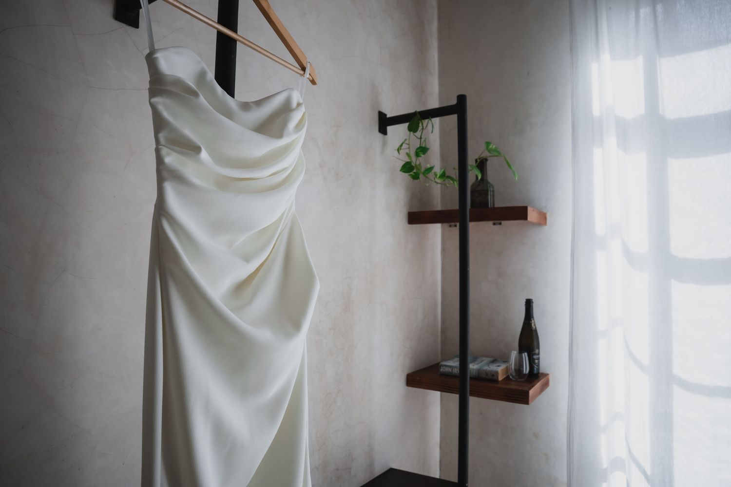 Elegant white satin wedding dress hanging on a wooden hanger near a shelf and window.