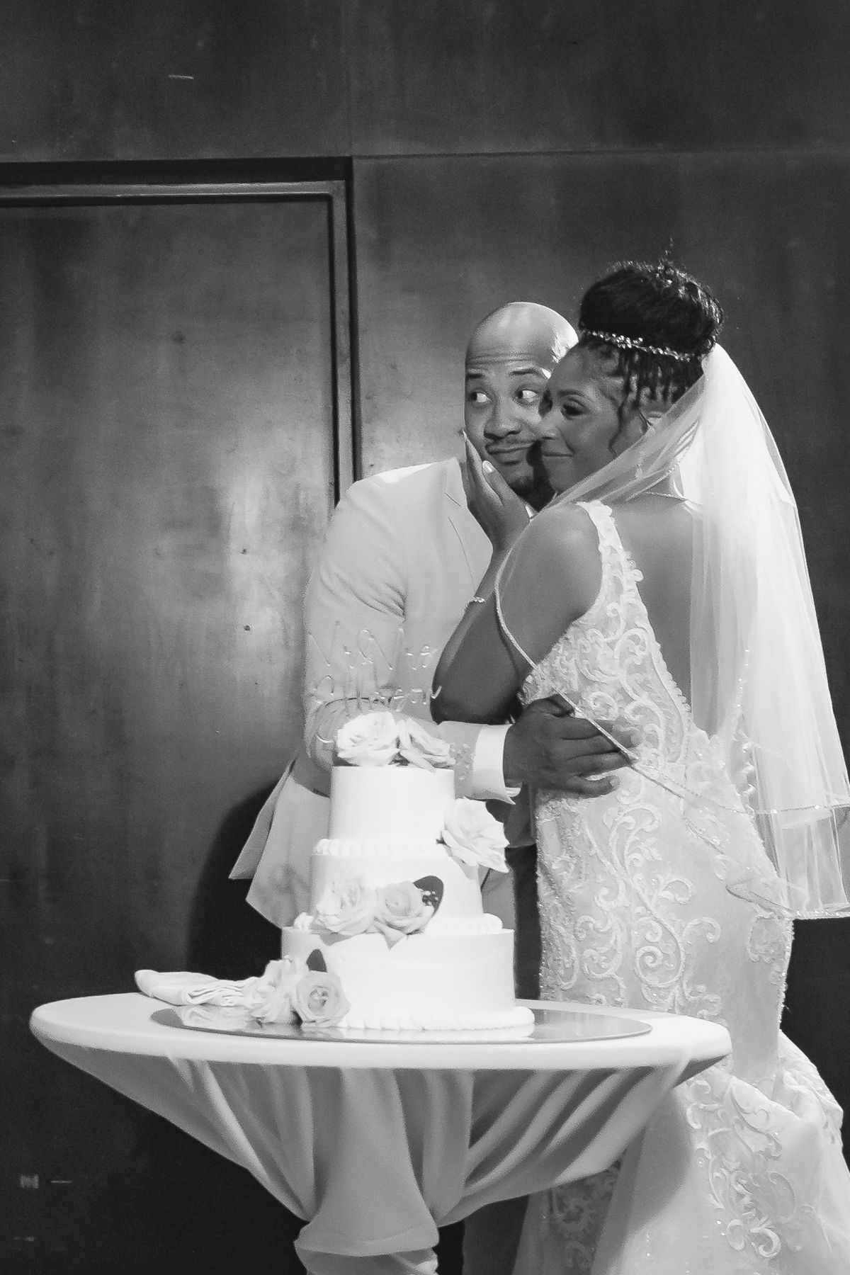 Black and white photo of a bride and groom sharing a tender moment beside their elegant tiered wedding cake.