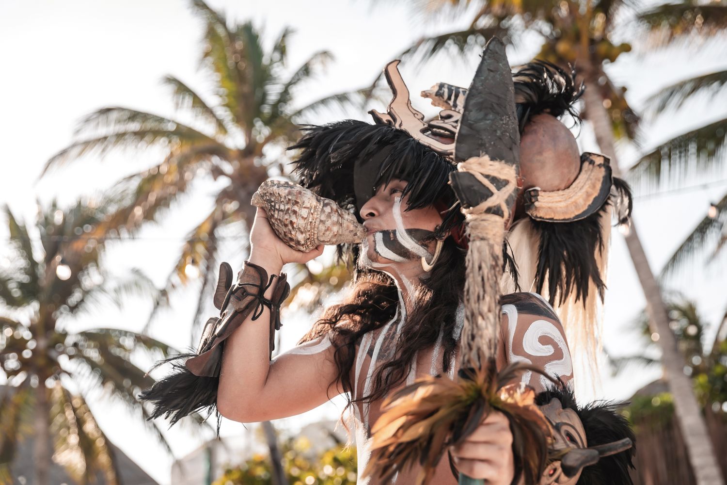 Tribal performer in elaborate mask and costume with palm trees in background at outdoor festival.
