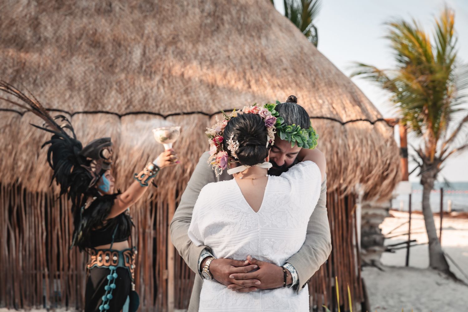 Couple embracing on beach near thatched hut during tropical wedding ceremony with costumed officiant.