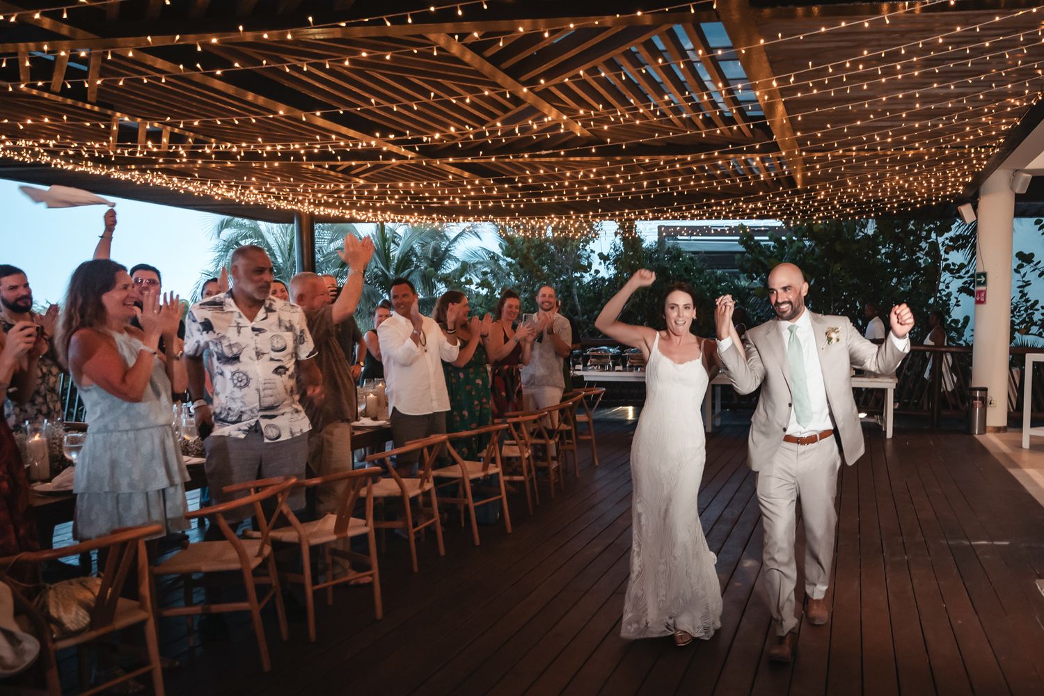 Newlywed couple makes grand entrance at outdoor wedding reception under string lights with cheering guests.