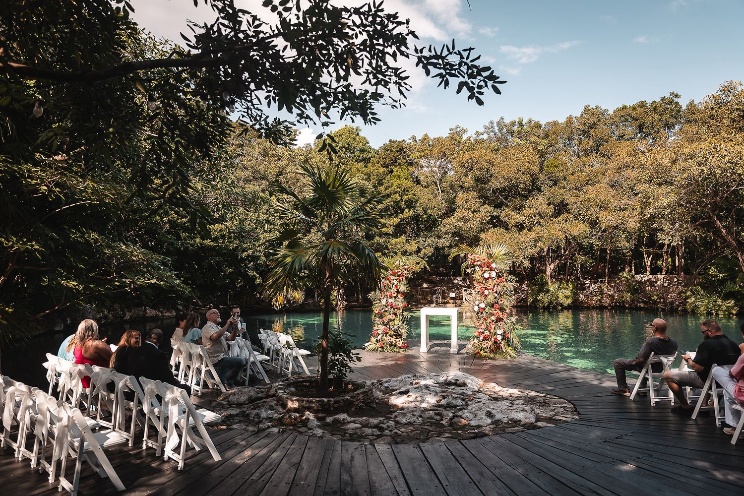 Outdoor wedding ceremony on wooden deck surrounded by lush jungle and turquoise water with white chairs.