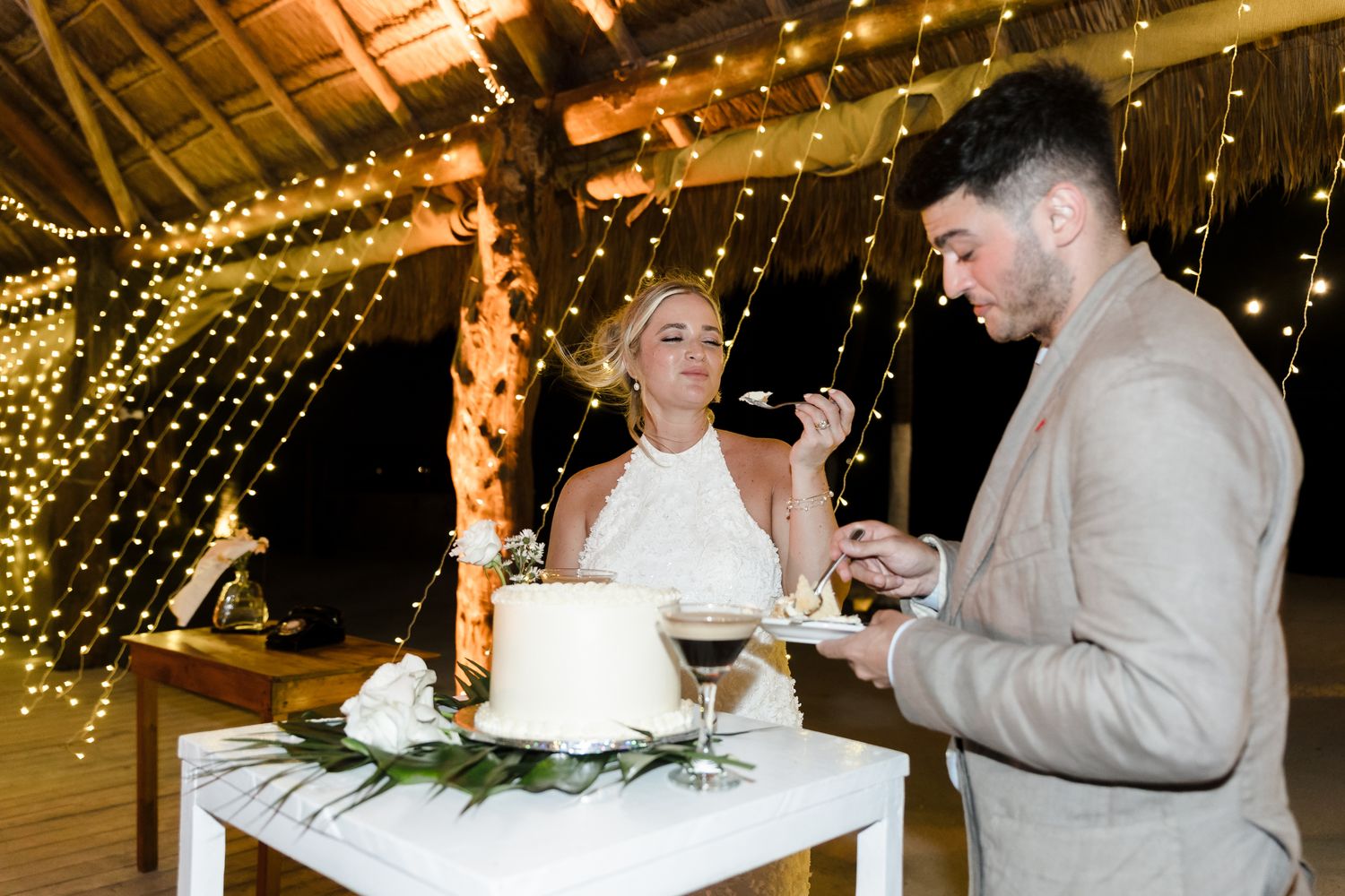 Bride and groom cut white wedding cake under fairy lights at tropical outdoor reception.