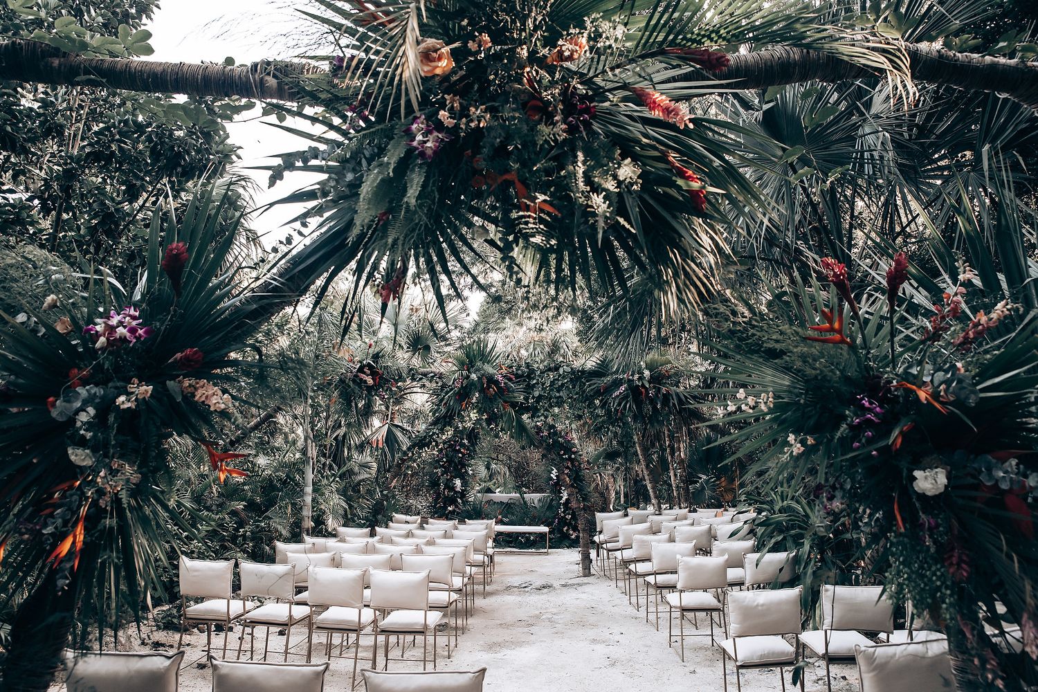 White chairs arranged in rows for an outdoor wedding ceremony under tropical palm trees and string lights.
