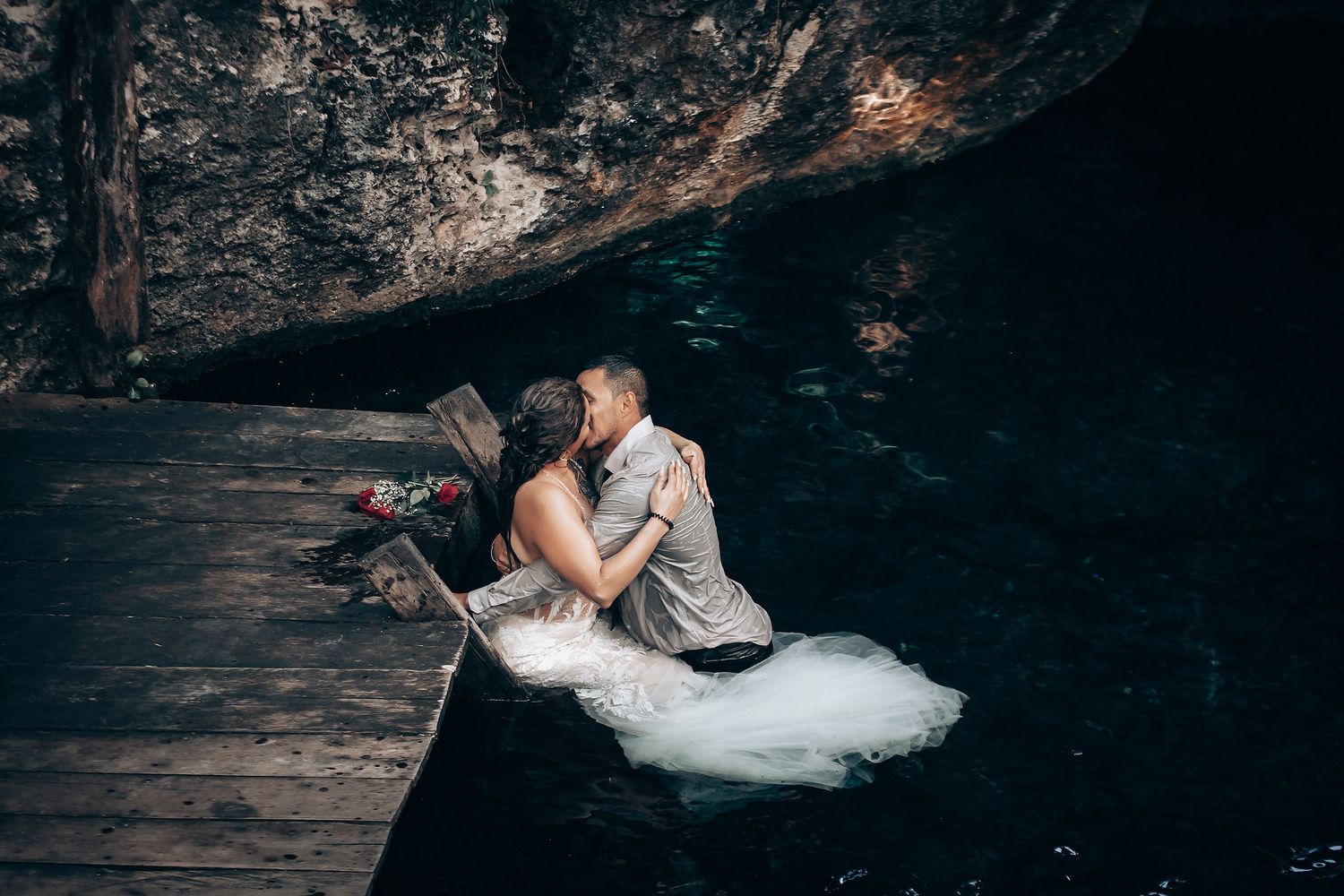 A couple in wedding attire poses on a rustic wooden platform in a forest setting.