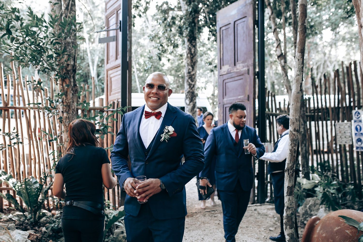 Wedding party gathers in an outdoor garden setting with bamboo fencing and trees in the background.