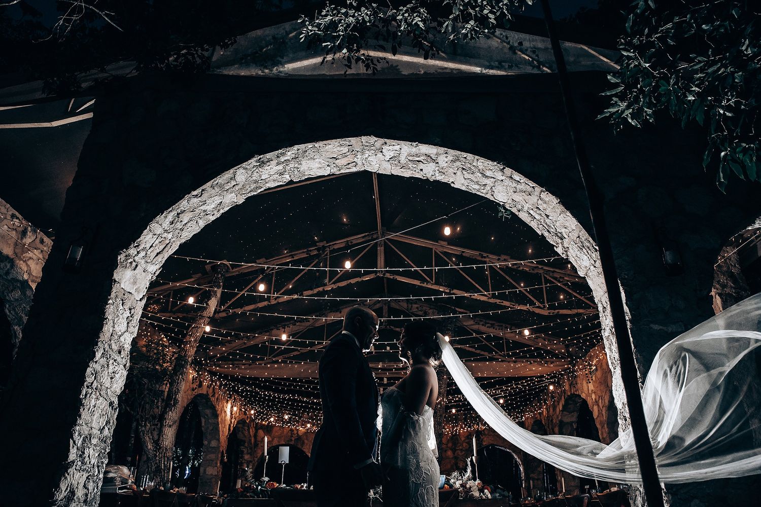 Wedding ceremony under illuminated stone arch with string lights and flowing fabric.