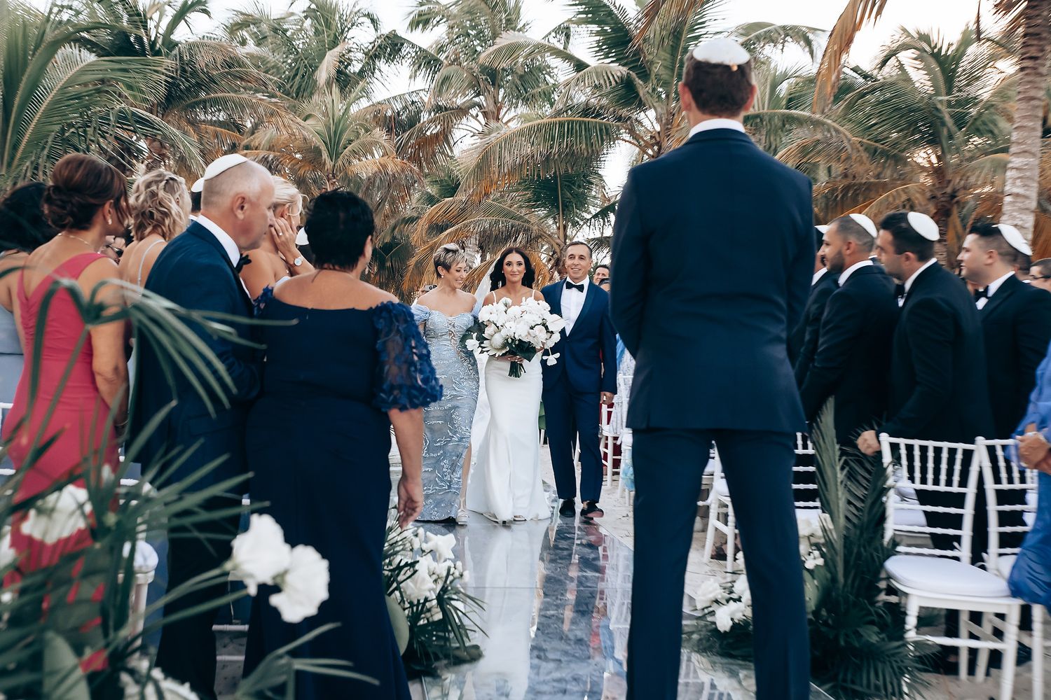 Wedding guests seated in white chairs during outdoor tropical ceremony.