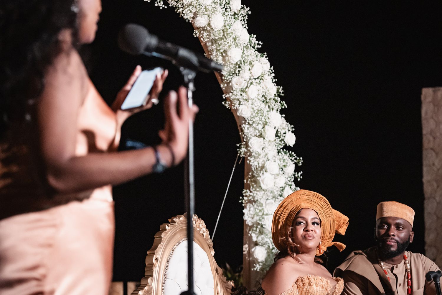Wedding guest speaks at microphone before seated couple under white floral arch at outdoor ceremony.