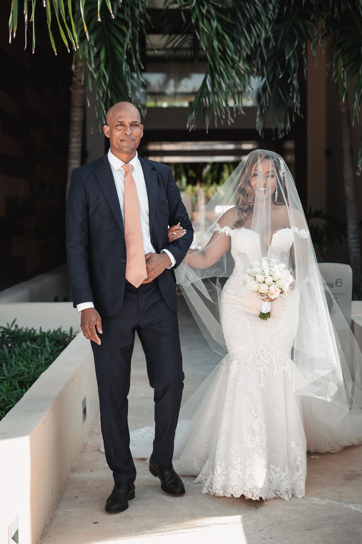 Bride in lace mermaid gown and veil walks with escort holding bouquet outdoors.