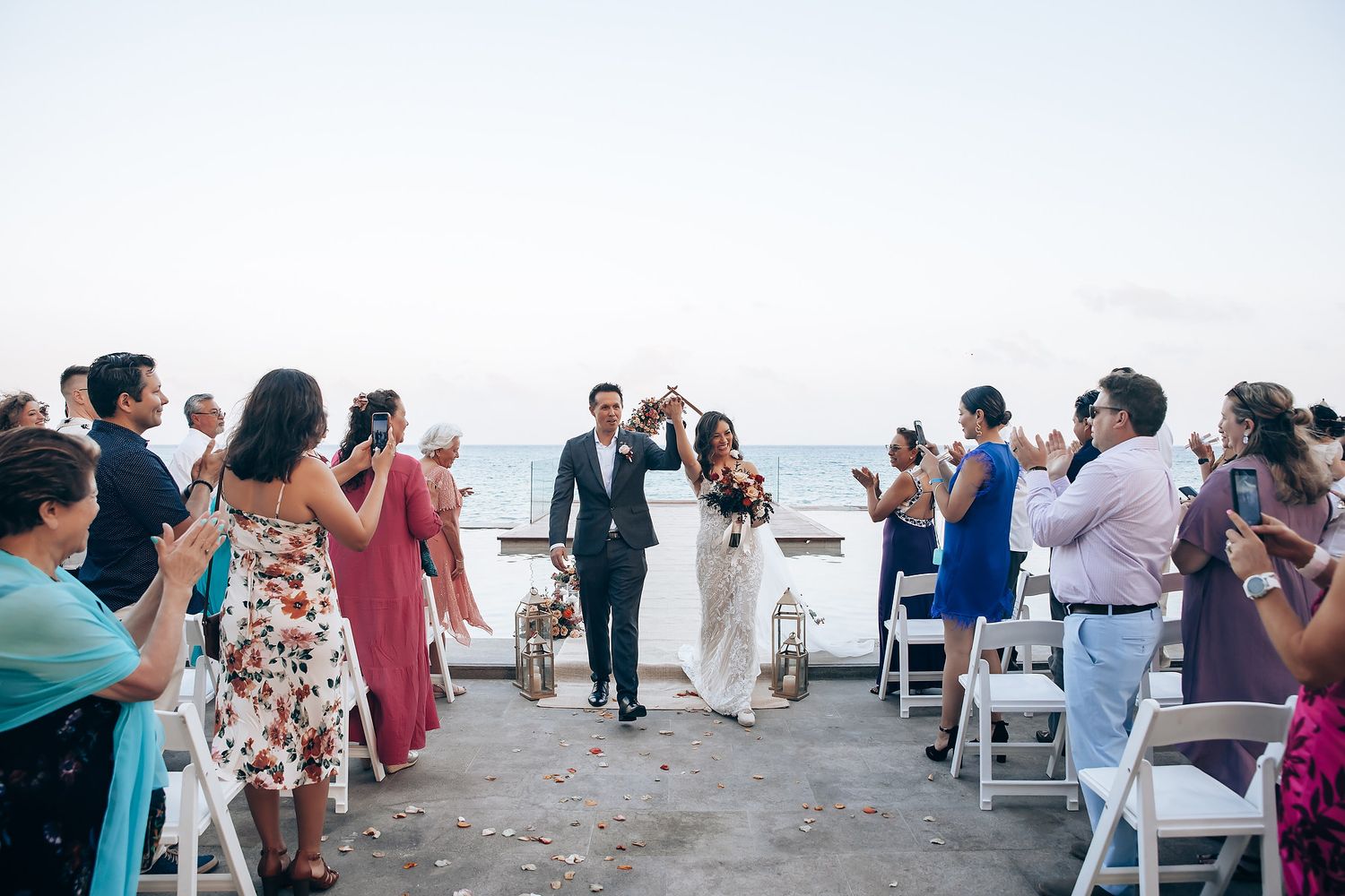 Panoramic view of beach wedding ceremony with guests seated on white chairs.