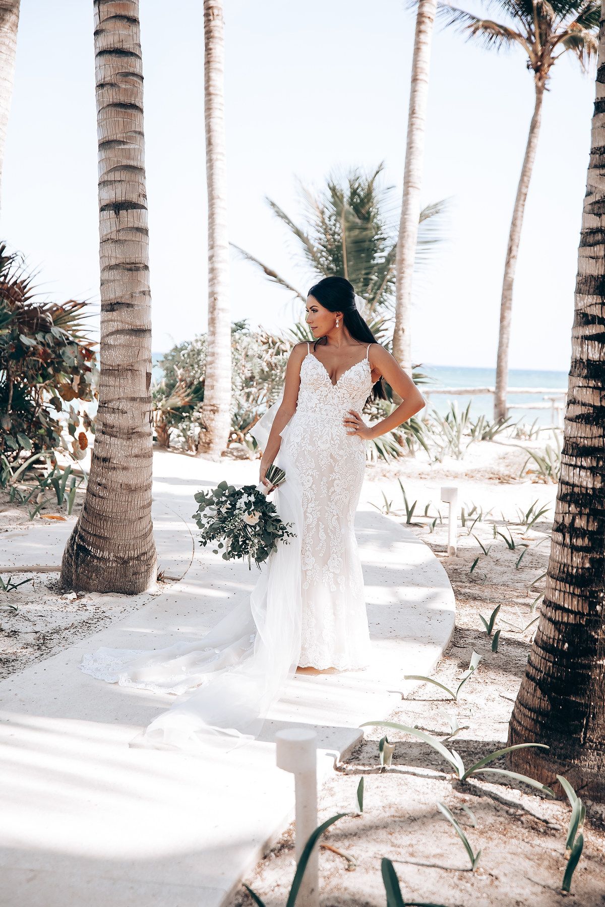 A white wedding dress flows in the breeze among palm trees on a tropical beach setting.
