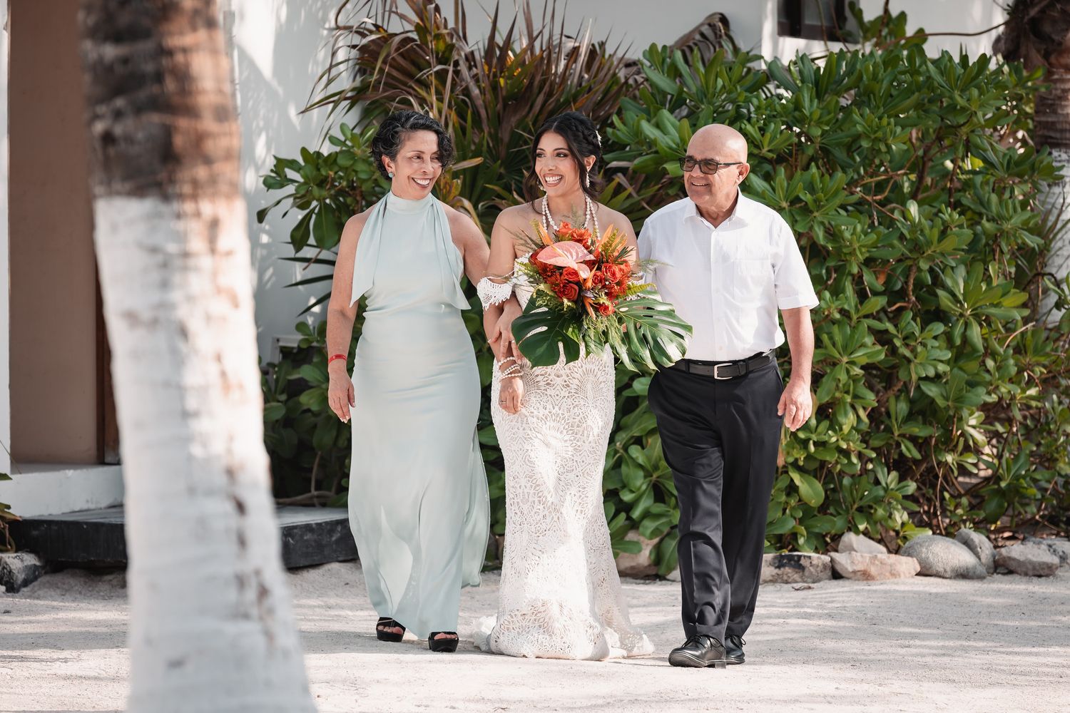 Bride in lace gown walks with parents toward beach wedding venue holding tropical floral bouquet.