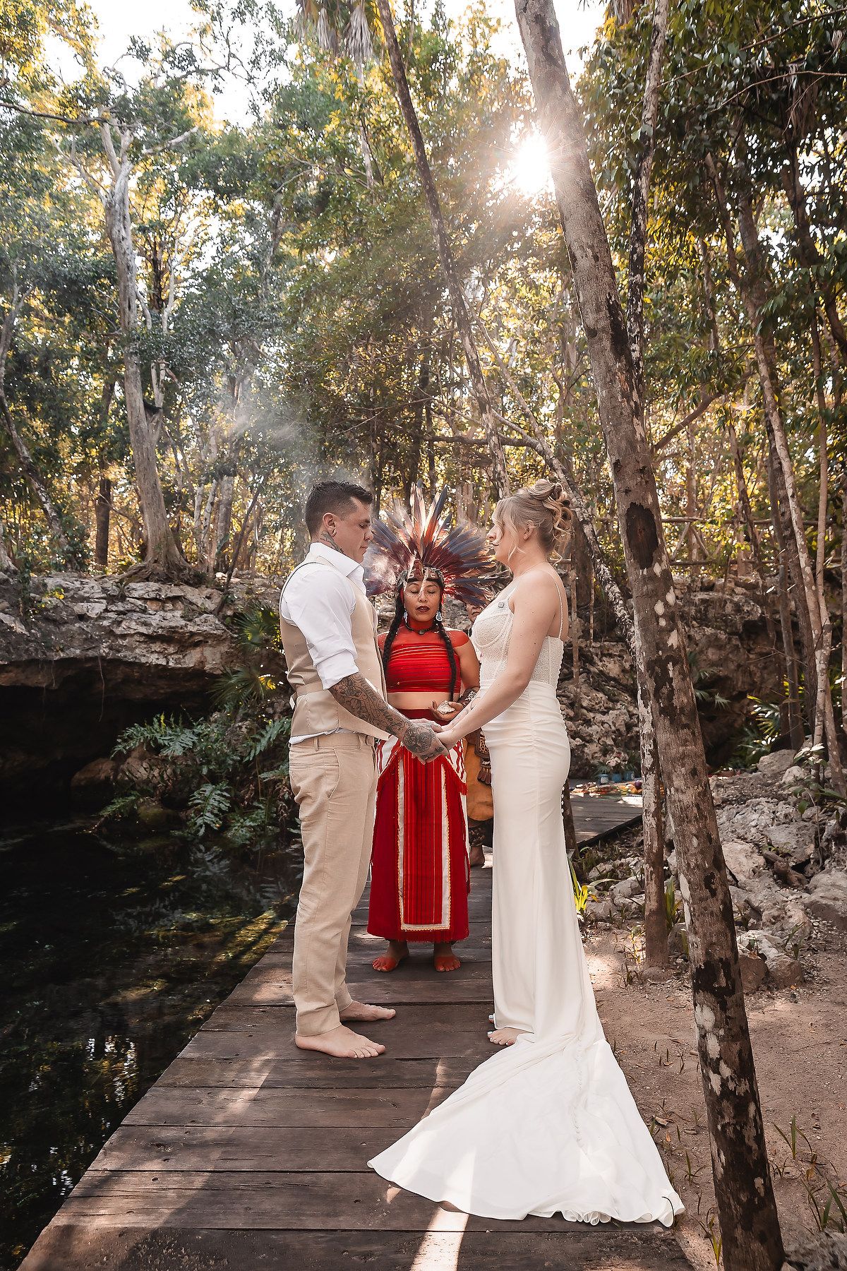 Couple exchanges vows in a jungle cenote ceremony officiated by a Mayan shaman in traditional red attire and headdress.