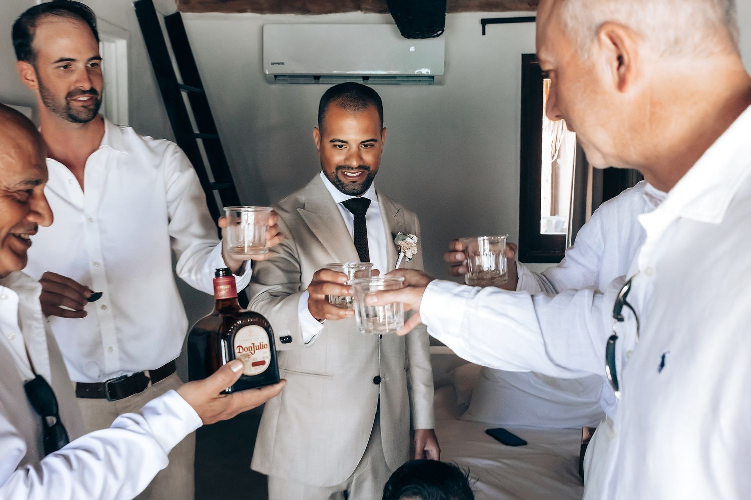Wedding party guests in white attire celebrate and toast with drinks at an indoor reception.