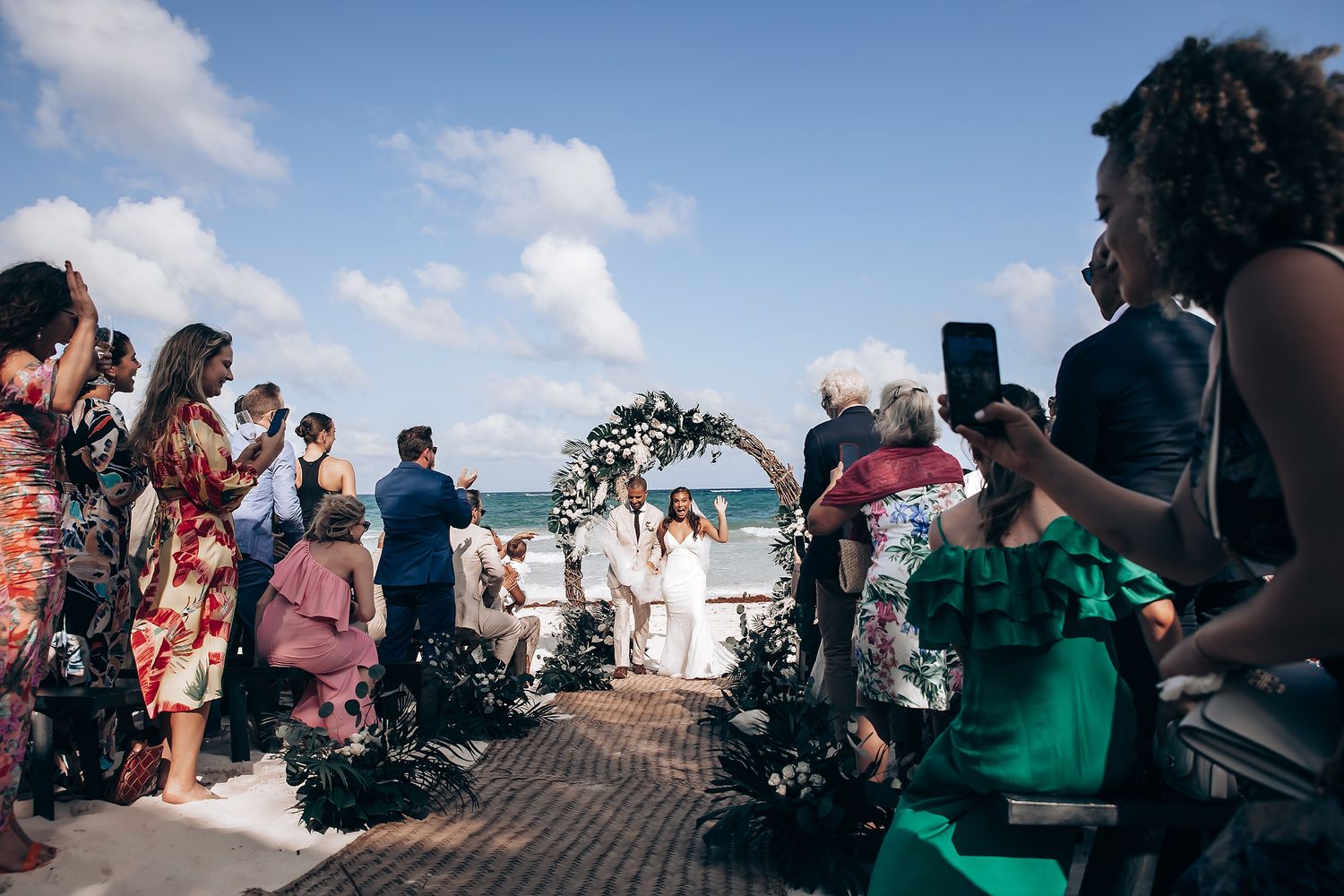 Wedding guests gathered around a beach ceremony setup with tropical decorations and ocean backdrop.