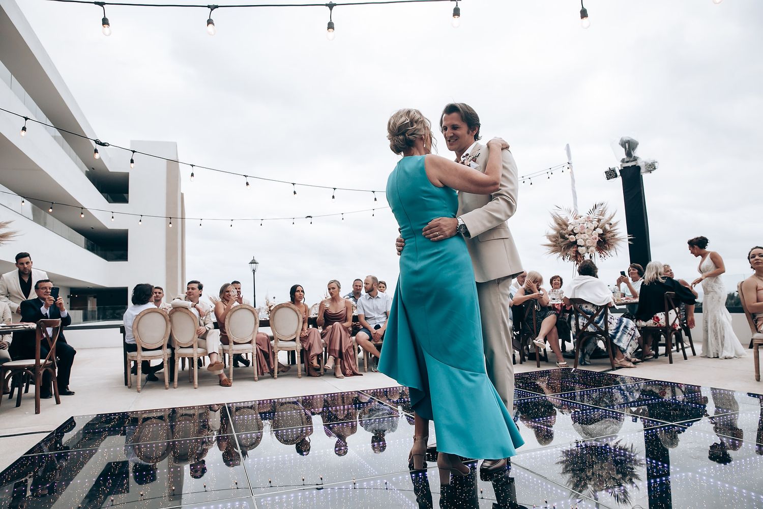 Couples dance together at an outdoor wedding reception under string lights at sunset.