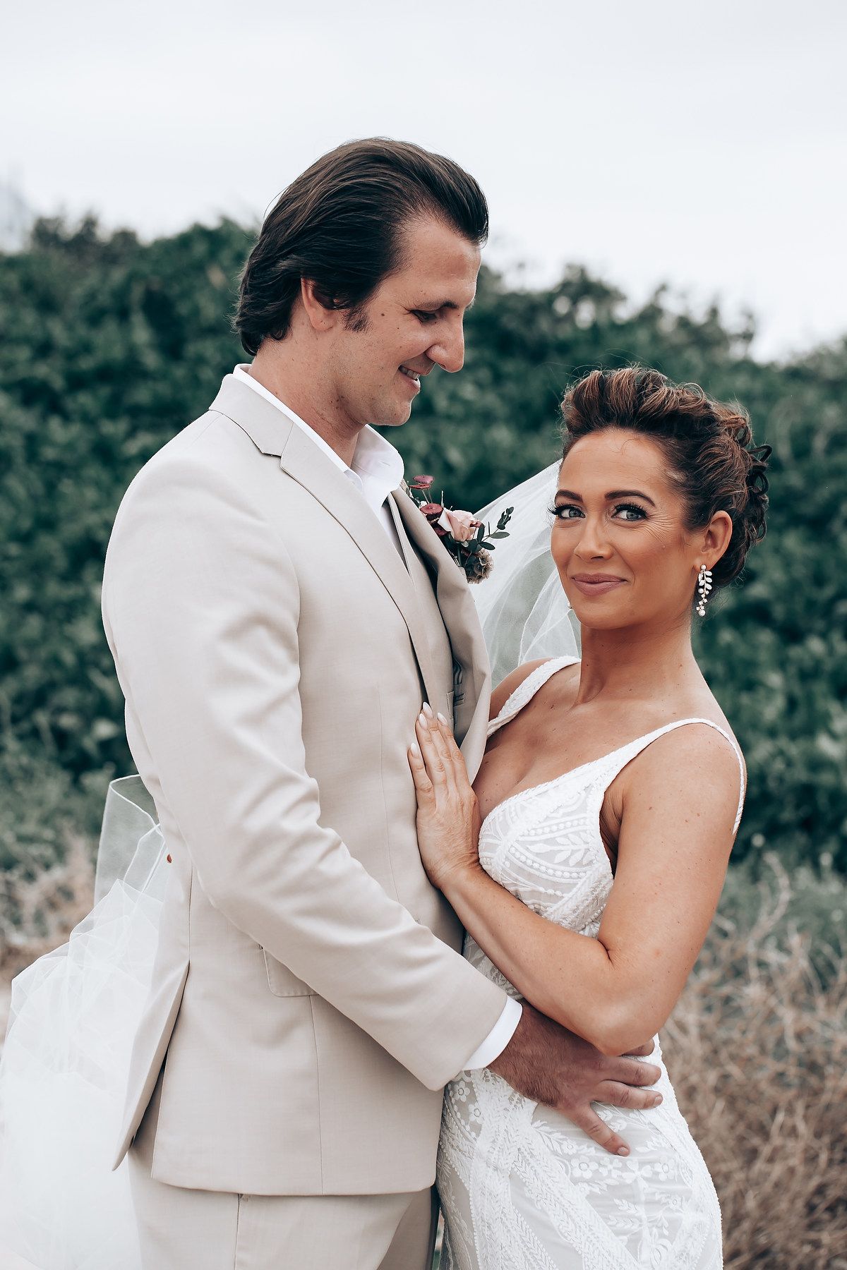 Romantic wedding couple embraces outdoors, with bride in white dress and groom in beige suit.