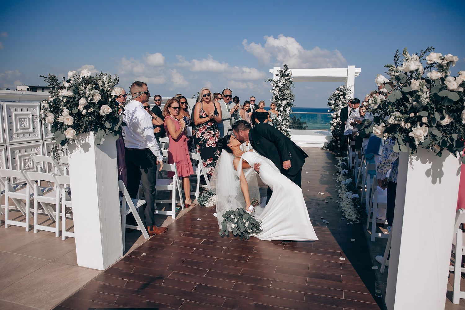 Beachfront wedding ceremony with white chairs and floral decorations along a wooden boardwalk.