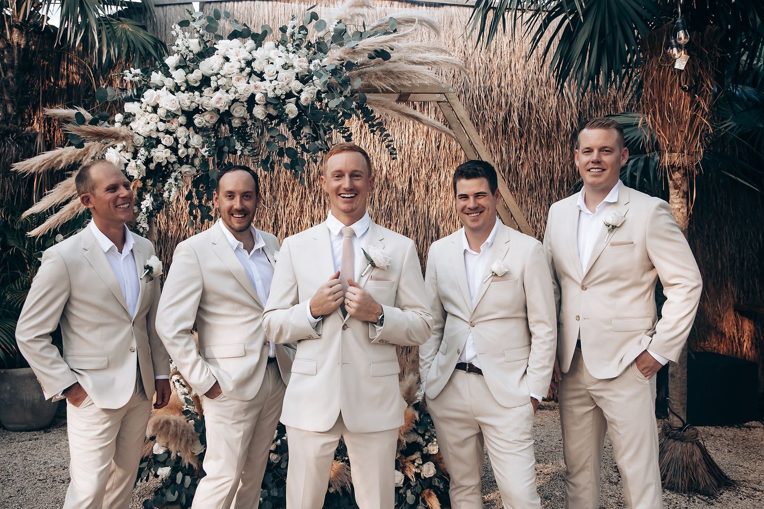 Groomsmen in crisp white suits pose together in front of a tropical floral wedding arch.