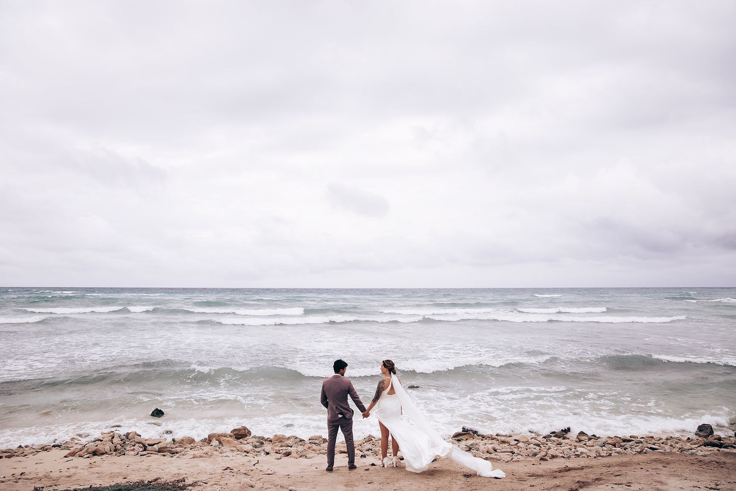 A couple walks along a rocky beach shoreline with waves crashing in the background.