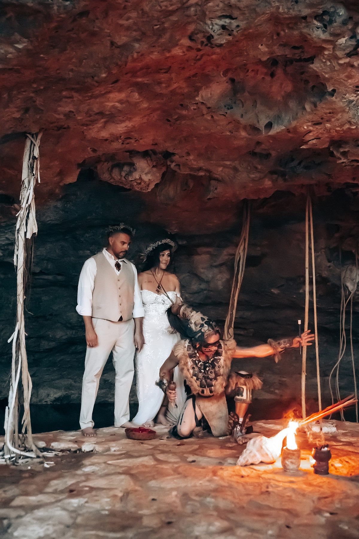 A romantic couple poses in a dimly lit cave with hanging vines and a warm fire glow creating atmospheric lighting.