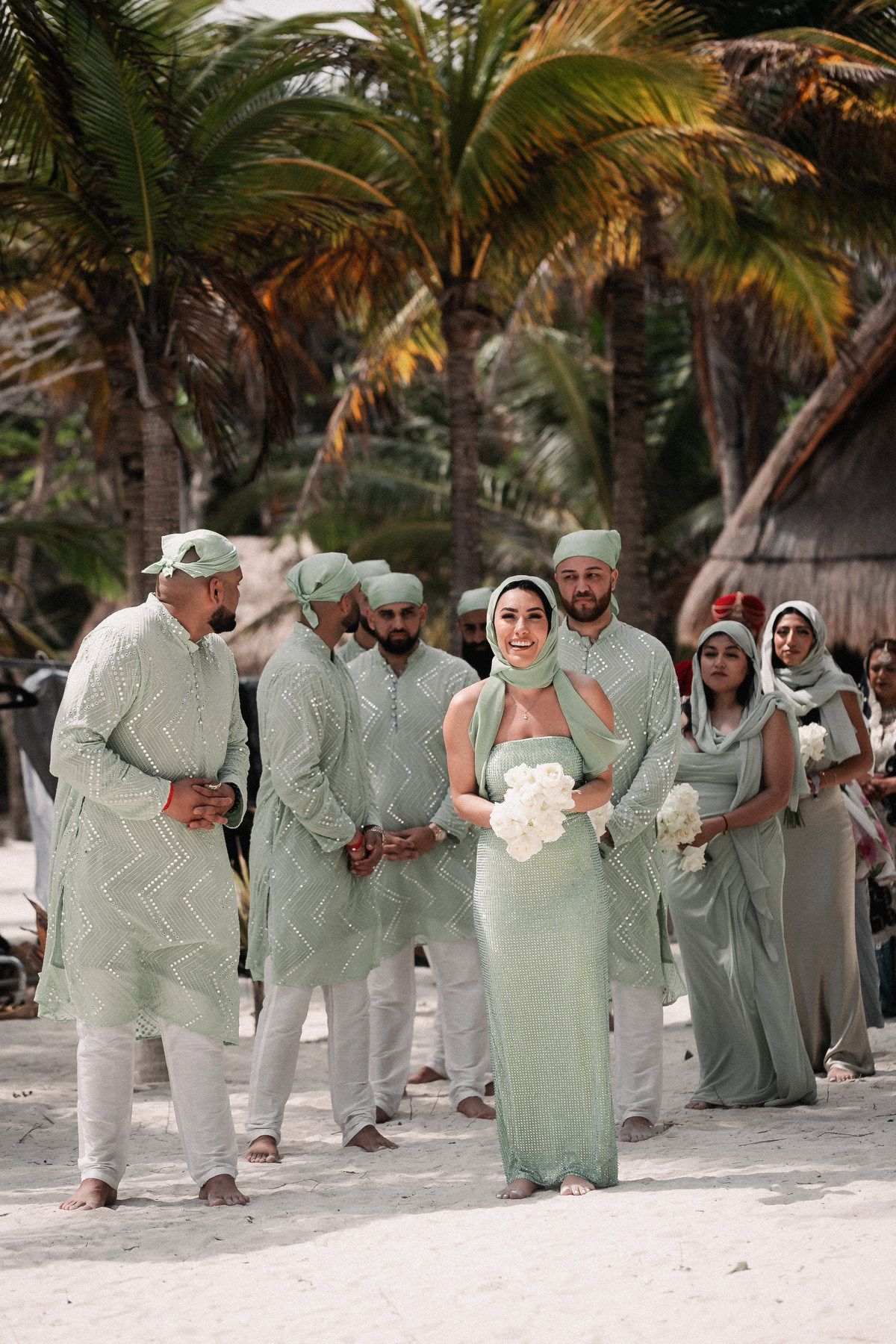 Wedding party in matching sage green outfits on a tropical beach with palm trees.