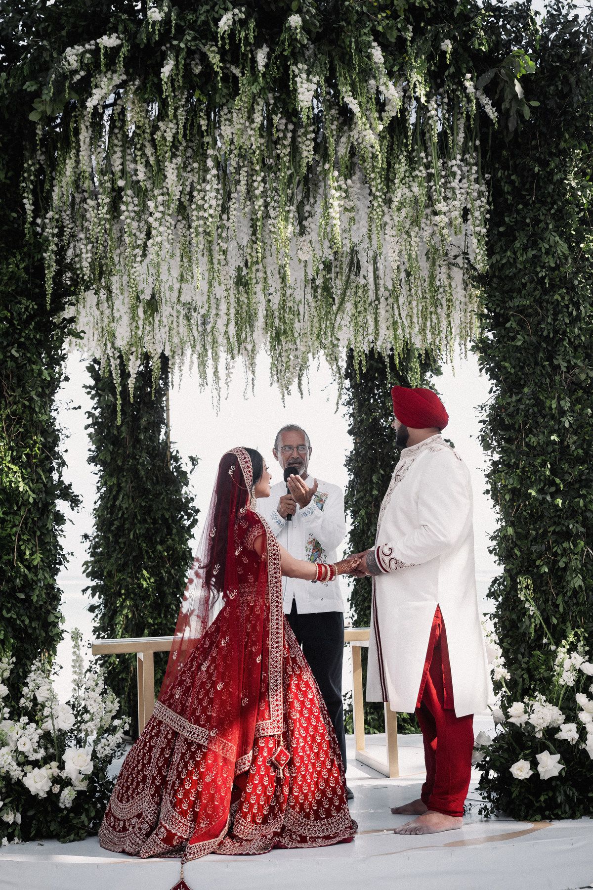 Sikh bride in red lehenga and groom in white sherwani exchange vows under lush green arch with white flowers.