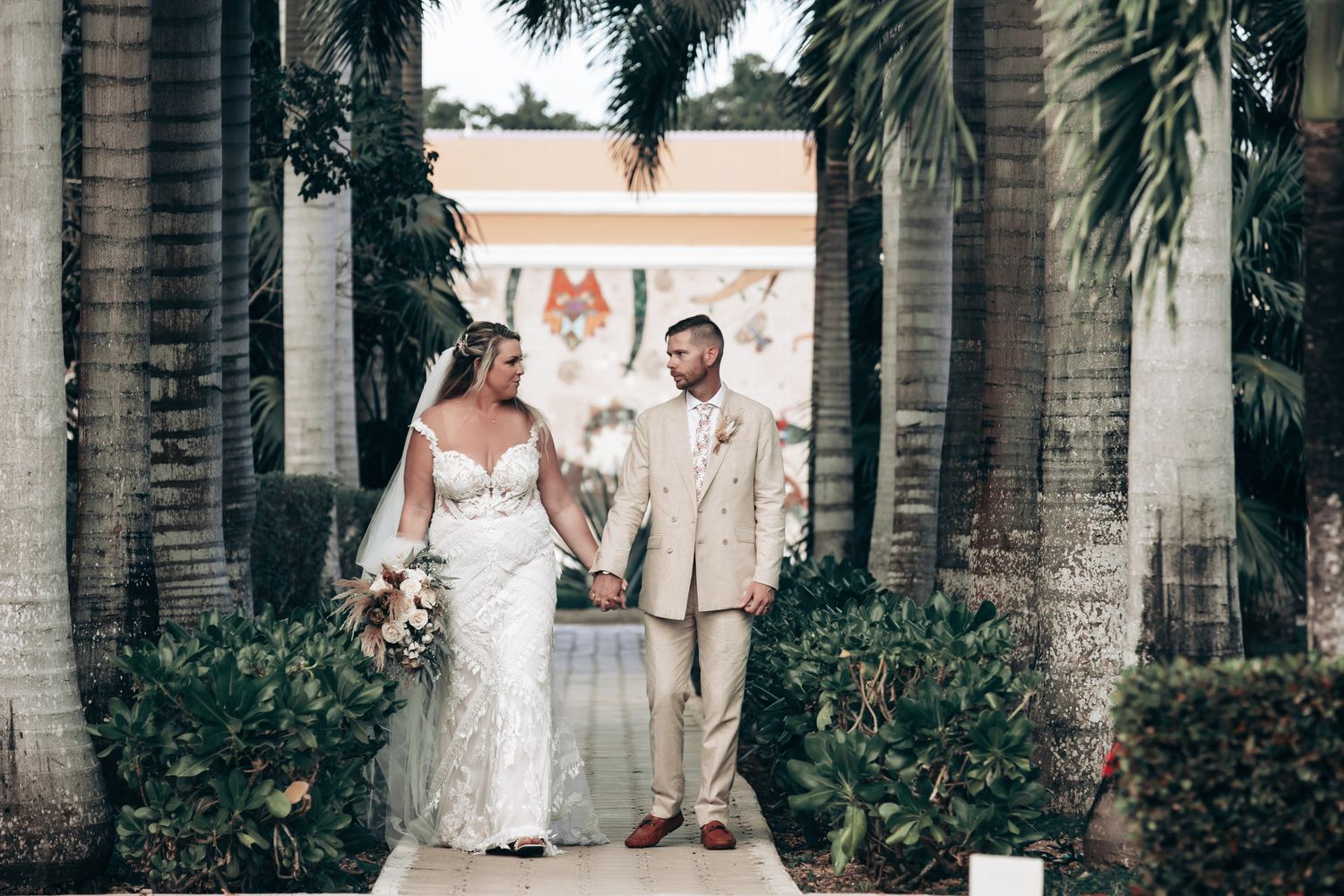Bride and groom holding hands walking through tropical palm tree lined path at outdoor wedding venue.