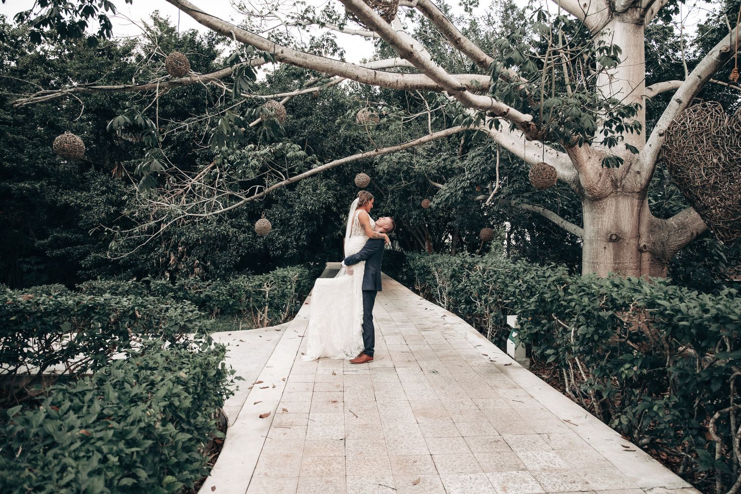 A bride and groom share a romantic embrace on a stone walkway surrounded by lush green trees and shrubs.