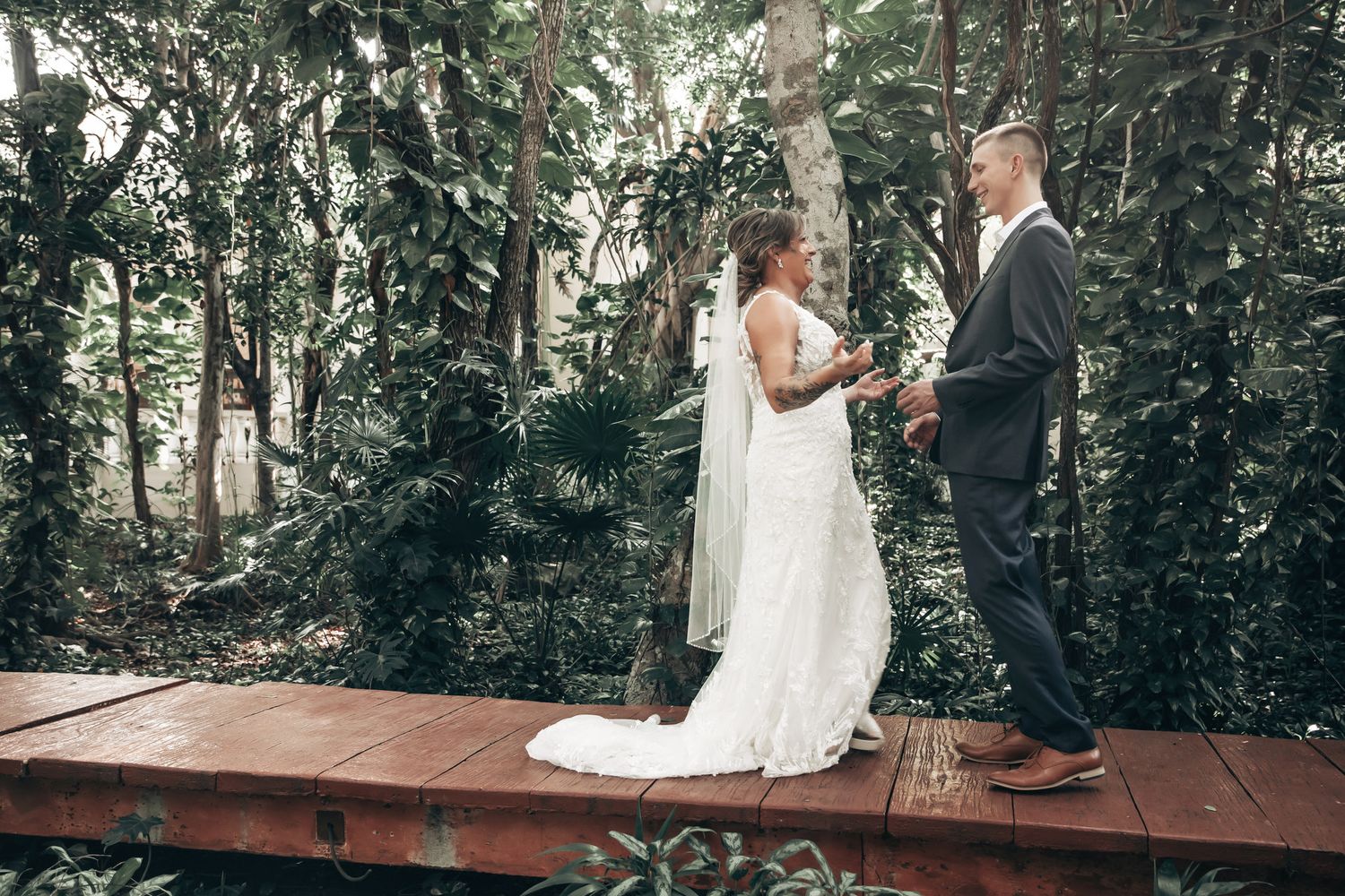 Bride and groom face each other on a rustic wooden platform amid dense green tropical trees.