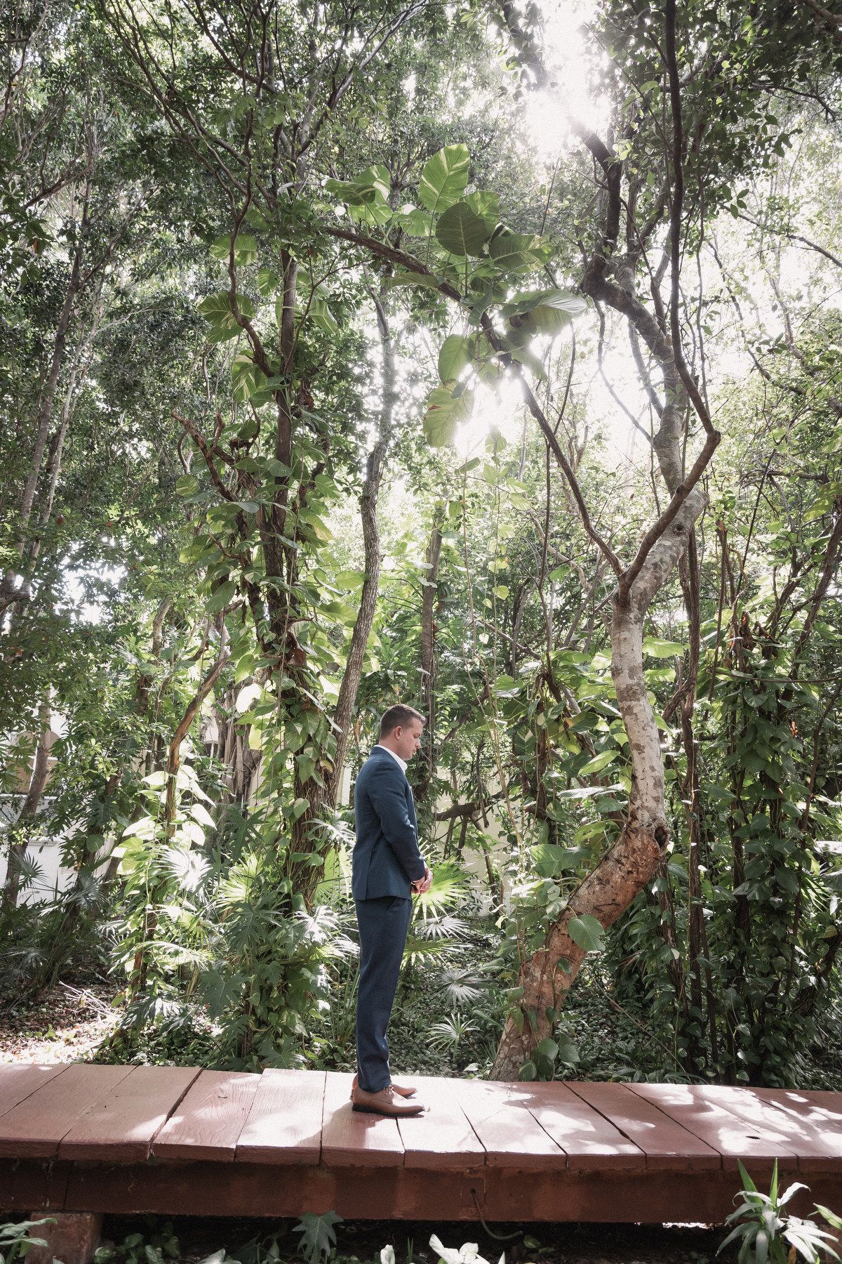 Groom stands alone on wooden deck surrounded by lush tropical forest before wedding ceremony.